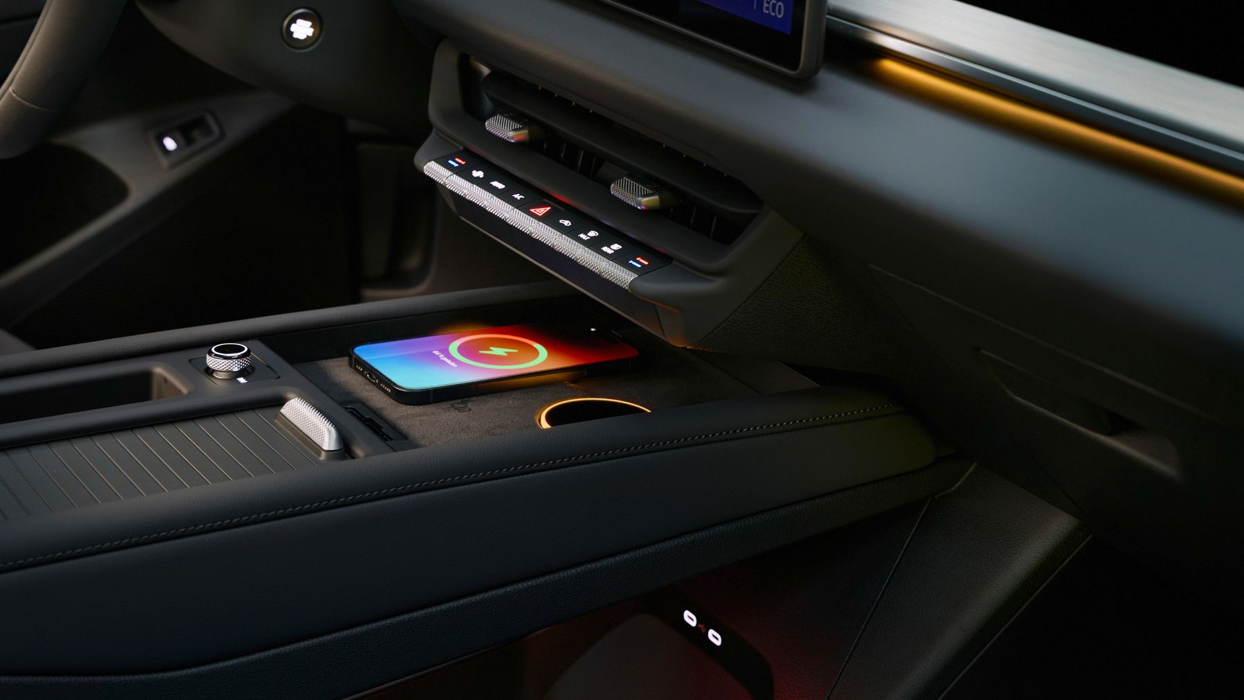 Nahaufnahme der Mittelkonsole eines modernen Autos mit beleuchtetem Ambientelicht. In der Ablagefläche liegt ein Smartphone, das kabellos geladen wird und ein buntes Ladesymbol anzeigt. Über der Ablage befinden sich Touch-Bedienelemente und ein großer, schwebend wirkender Bildschirm.