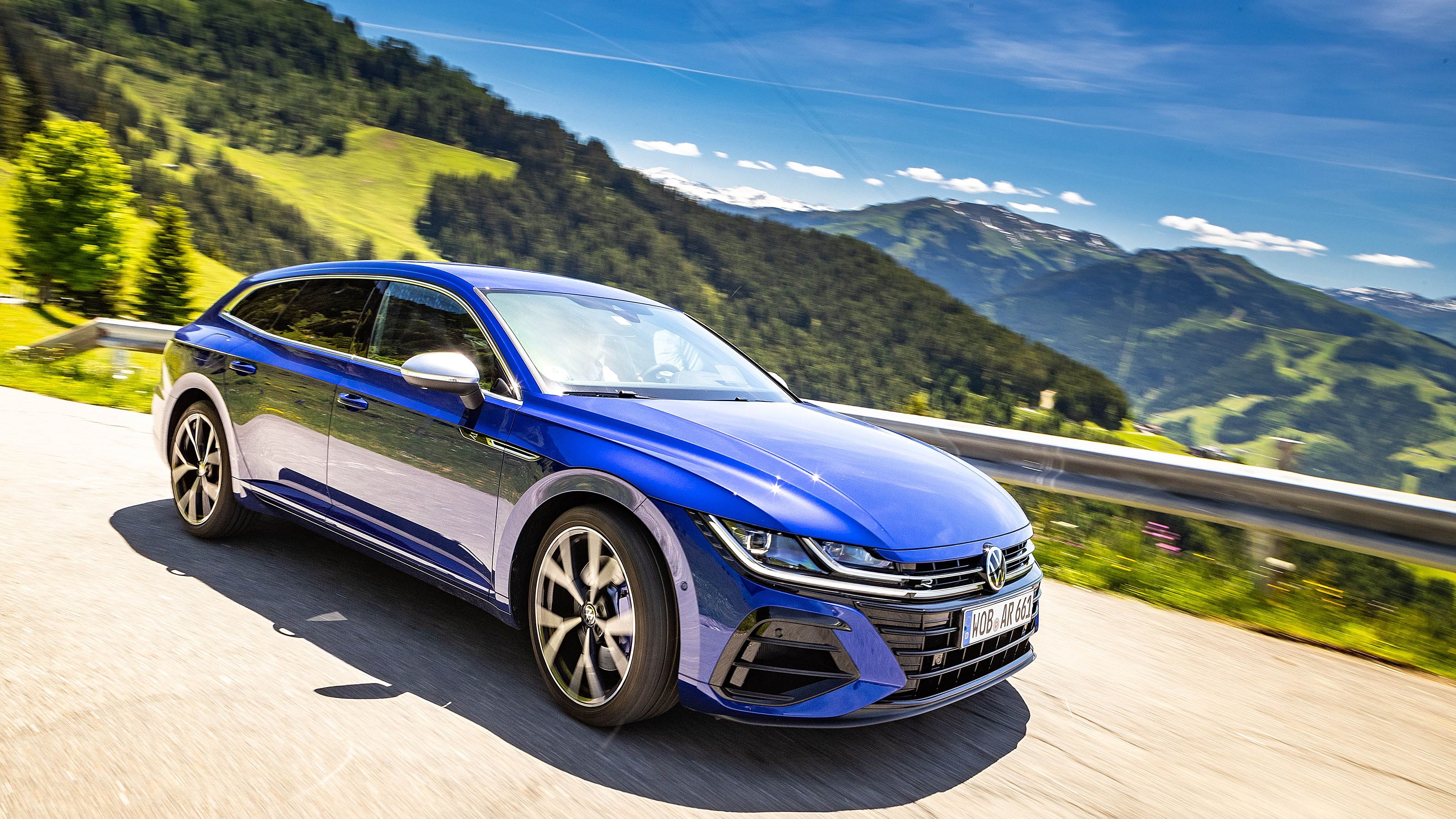 Ein blaues VW Arteon R Shooting brake fährt auf einer malerischen Bergstraße an einem sonnigen Tag. Die umliegende Landschaft ist grün und hügelig. Der Himmel ist blau mit wenigen Wolken.