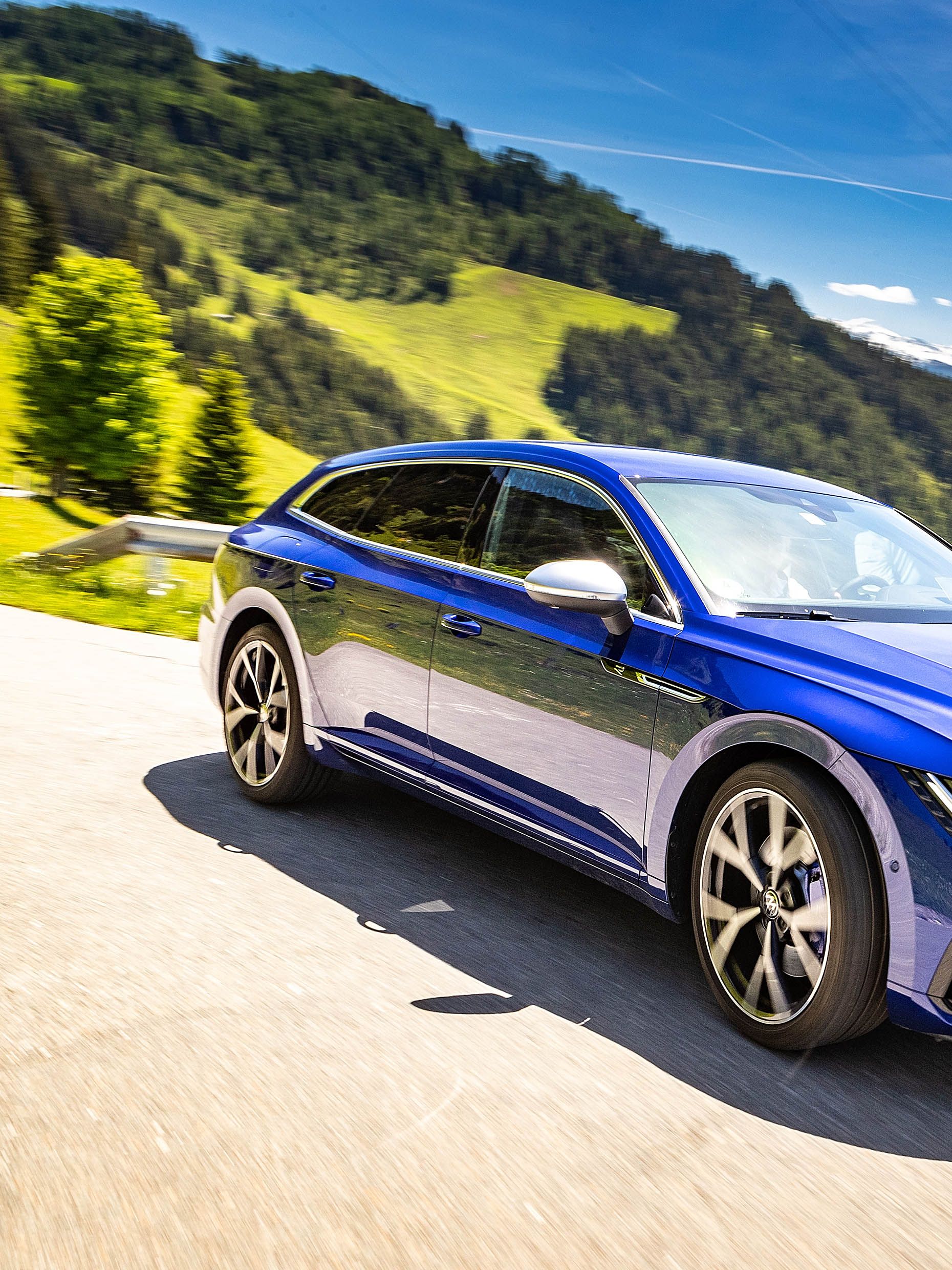 Ein blaues VW Arteon R Shooting brake fährt auf einer malerischen Bergstraße an einem sonnigen Tag. Die umliegende Landschaft ist grün und hügelig. Der Himmel ist blau mit wenigen Wolken.