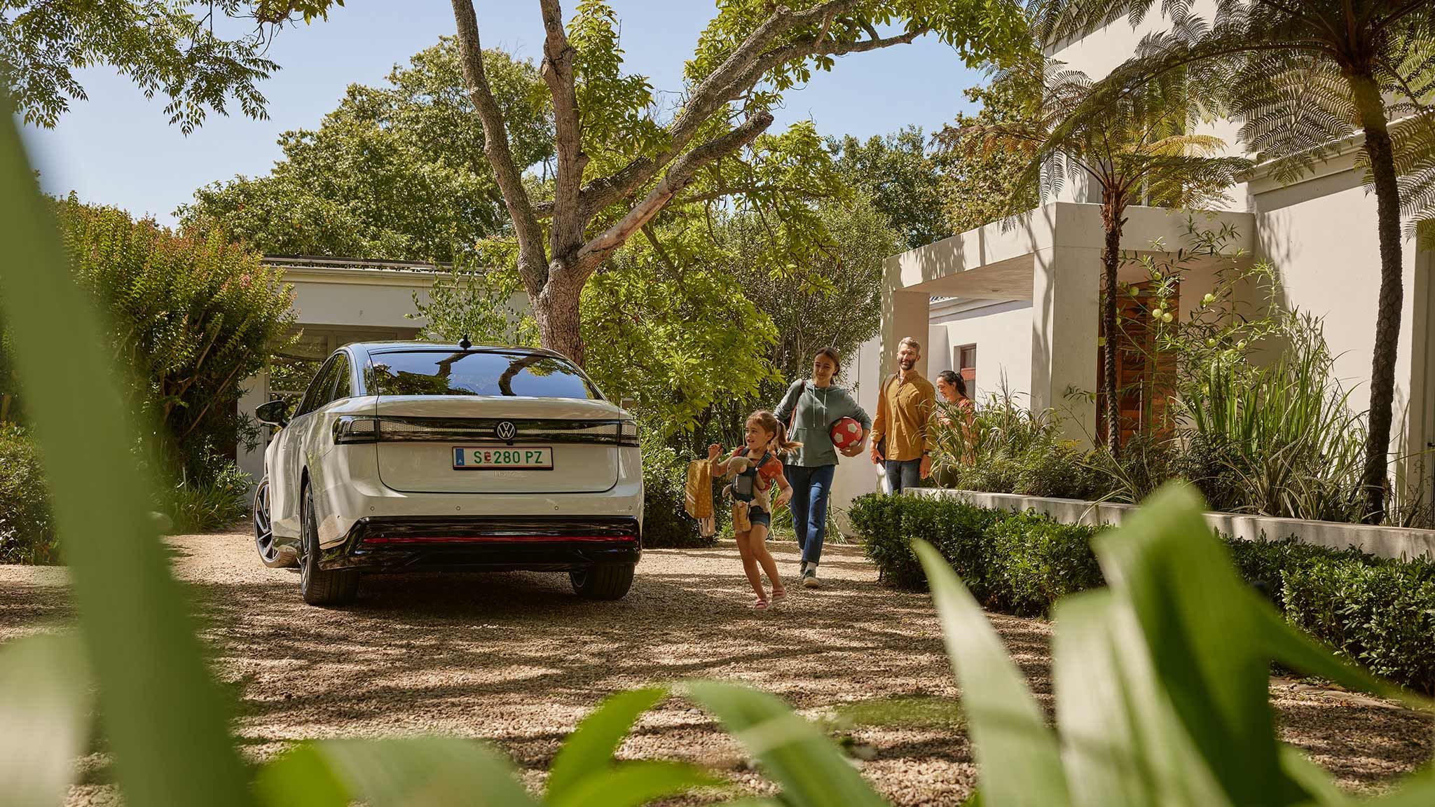 Eine Familie kommt gemeinsam mit einem Hund auf einem Grundstück an. Im Vordergrund sieht man ein Elektroauto, das in der Einfahrt geparkt ist. Die Umgebung ist grün und gut gepflegt.