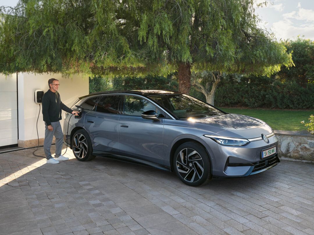 Ein ID.7 Tourer steht in einer Einfahrt vor einem Haus, ein Mann steckt den Ladestecker in die Ladebuchse. 