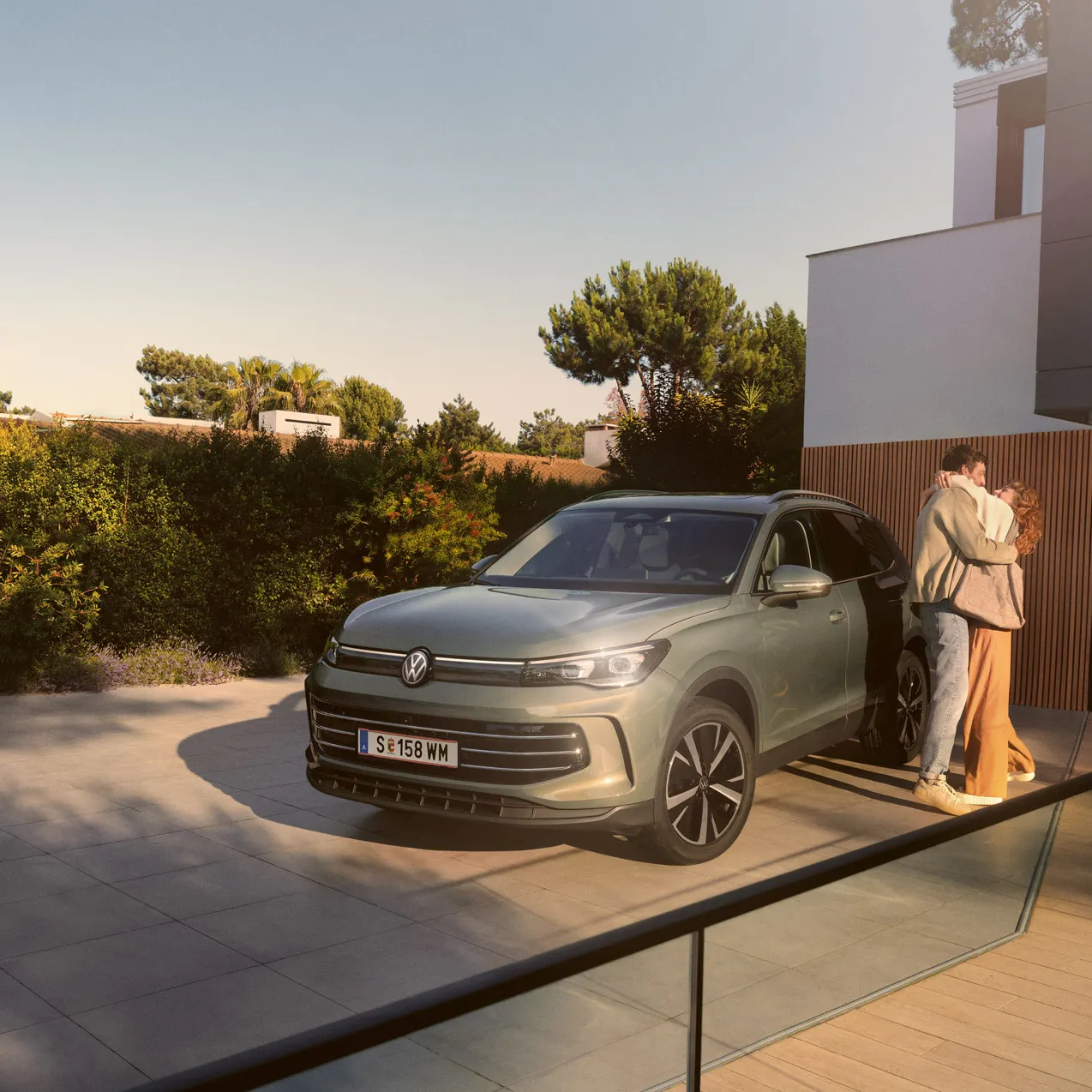 Eine Frau und ein Mann umarmen sich, neben ihnen steht ein VW Tiguan Elegance in der Farbe Cipressino-Grün Metallic