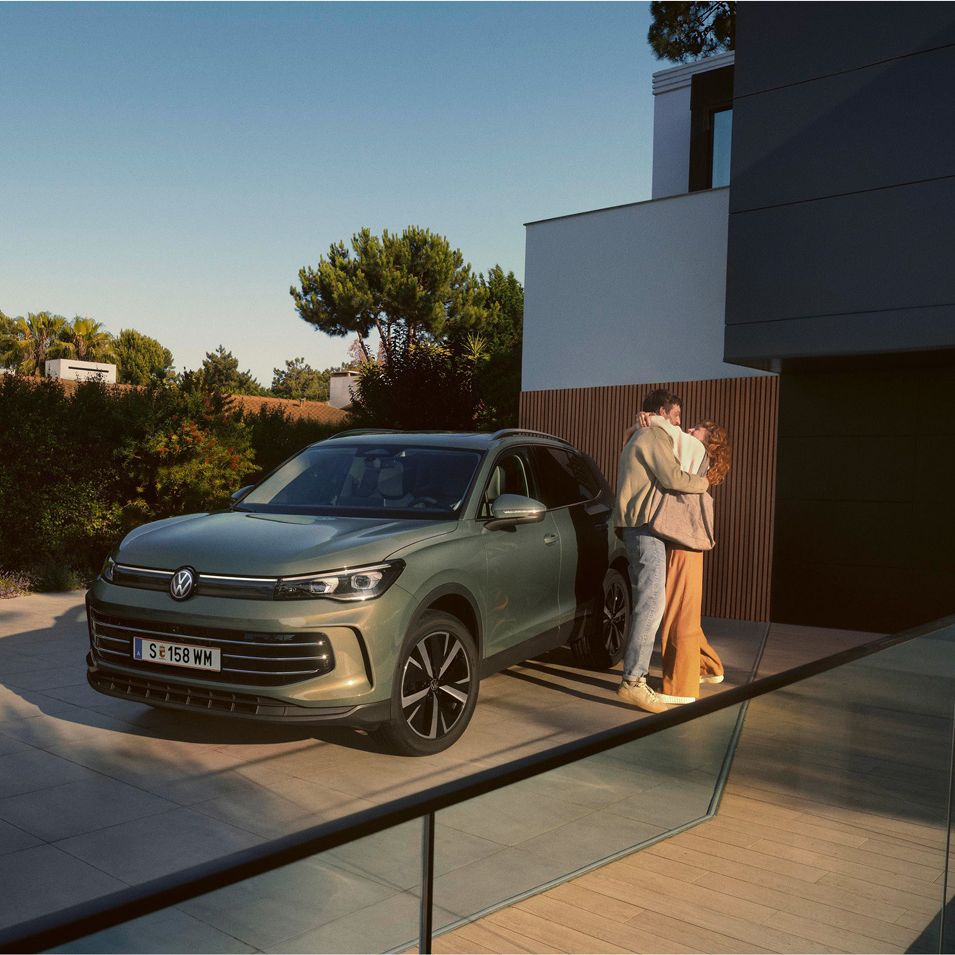 Eine Frau und ein Mann umarmen sich, neben ihnen steht ein VW Tiguan Elegance in der Farbe Cipressino-Grün Metallic