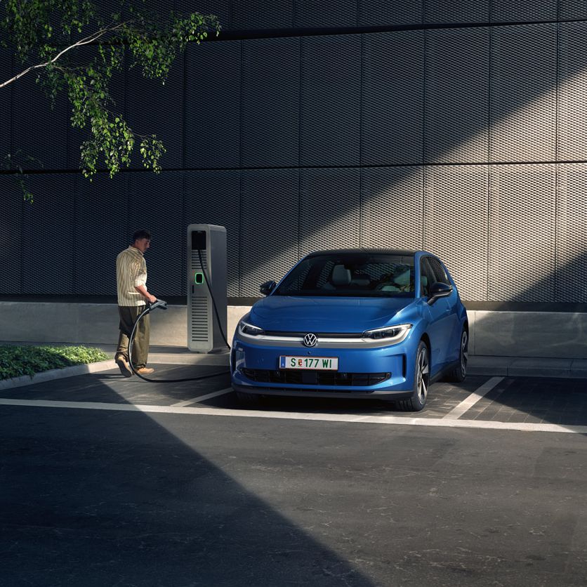 Ein blauer ID. Polo steht auf einem Parkplatz neben einer Ladesäule. Im Hintergrund sind ein Gebäude und ein Baum zu sehen.