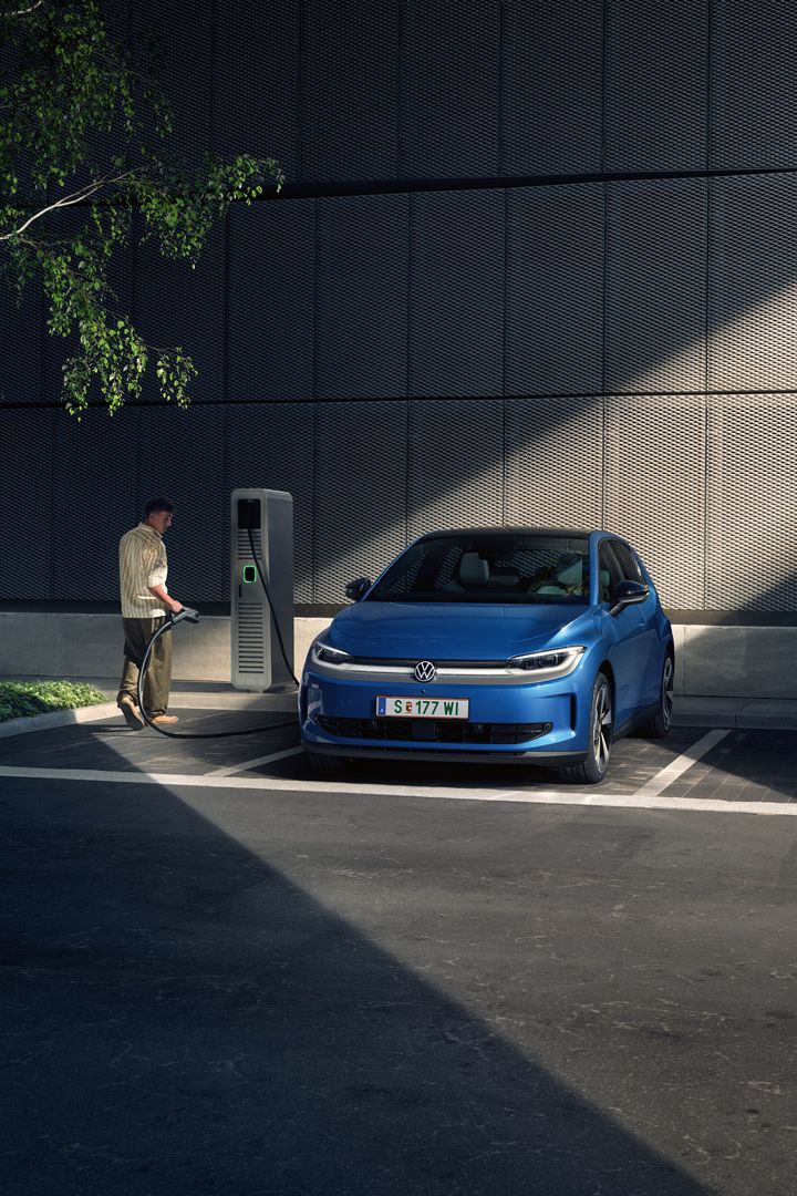 Ein blauer ID. Polo steht auf einem Parkplatz neben einer Ladesäule. Im Hintergrund sind ein Gebäude und ein Baum zu sehen.