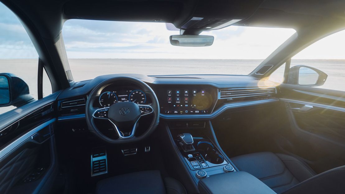 Innenraum eines Touareg R mit Lederpolstern und digitalem Armaturenbrett. Durch die Fenster sieht man eine helle Landschaft. Die Steuerungselemente sind beleuchtet und gut sichtbar.