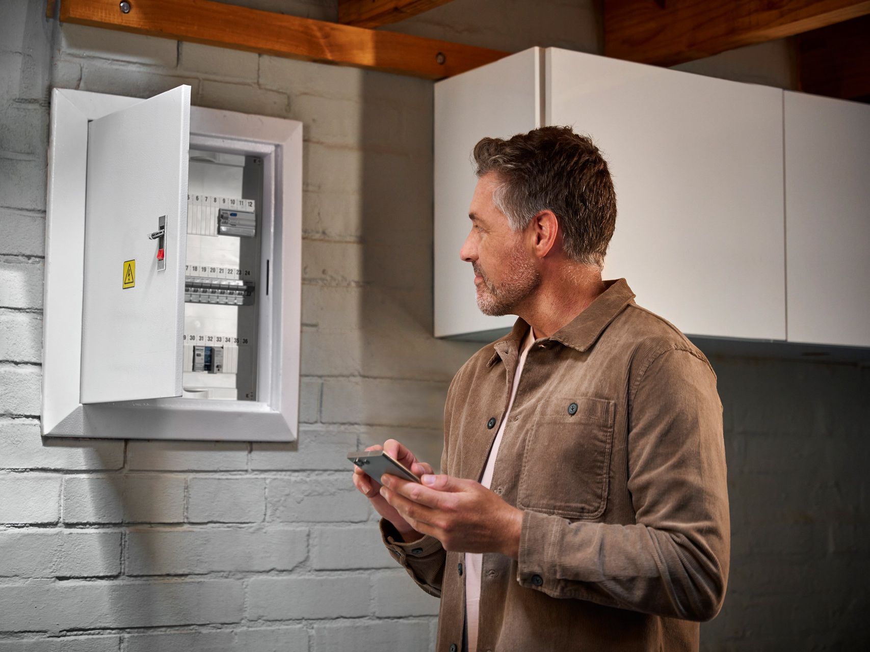 Ein Mann mittleren Alters steht in einer Garage oder Werkstatt vor einem geöffneten Sicherungskasten in der Wand. Er hält ein Smartphone in der Hand und blickt prüfend auf die Sicherungen. Im Hintergrund sind weiße Hängeschränke und eine gemauerte Wand zu sehen.
