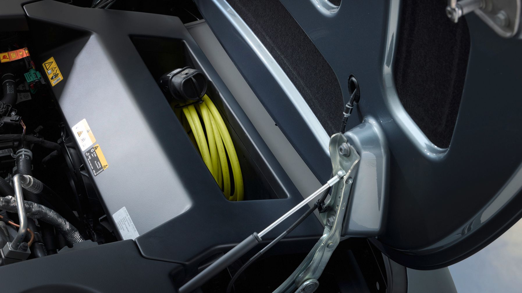 Detailansicht eines geöffneten Ladefachs an einem Elektrofahrzeug, in dem ein gelbes Ladekabel sauber aufgewickelt liegt. Sichtbar sind Gasdruckdämpfer, Scharniere und verschiedene technische Komponenten sowie Sicherheitsaufkleber.
