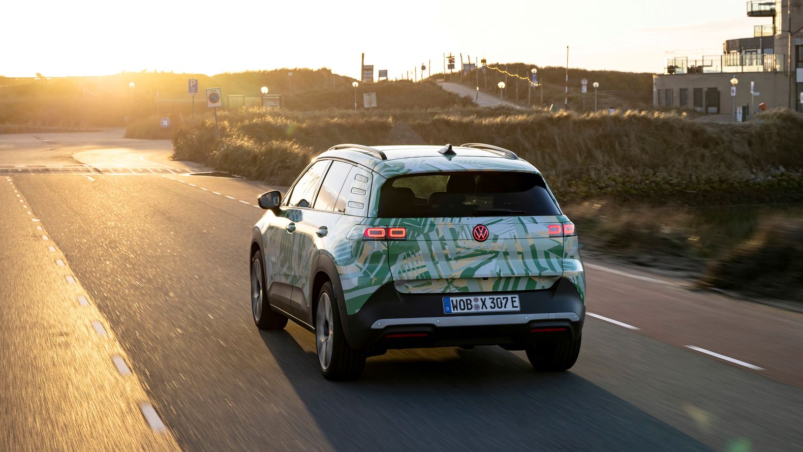 Ein Auto fährt auf einer Straße in einer Landschaft bei Sonnenuntergang. Das Fahrzeug hat ein grünes und weißes Design und ist ein elektrisches Modell. Im Hintergrund sind Gebäude und Hügel zu sehen.