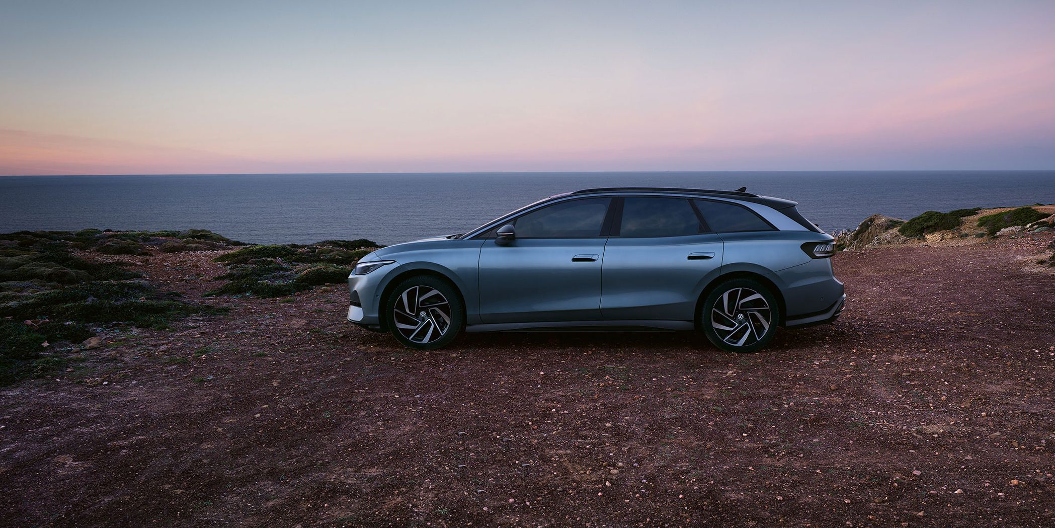 Seitenansicht des VW ID.7 in der Farbe Stonewashed Blue geparkt an der Küste mit Sonnenuntergang