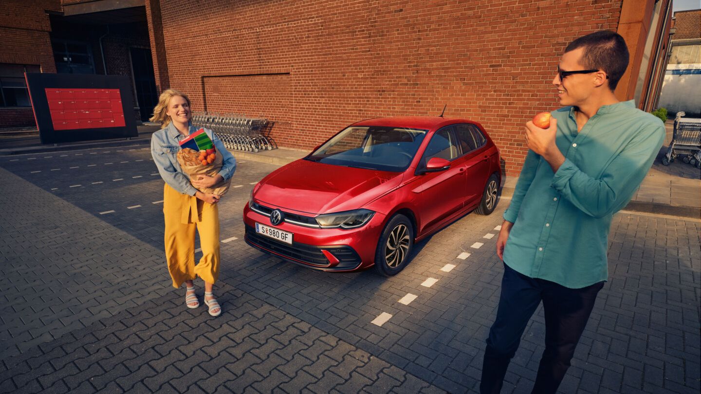 Ein roter VW Polo steht auf einem Parkplatz vor einem Supermarkt, ein lachendes Paar steht mit Einkäufen daneben.