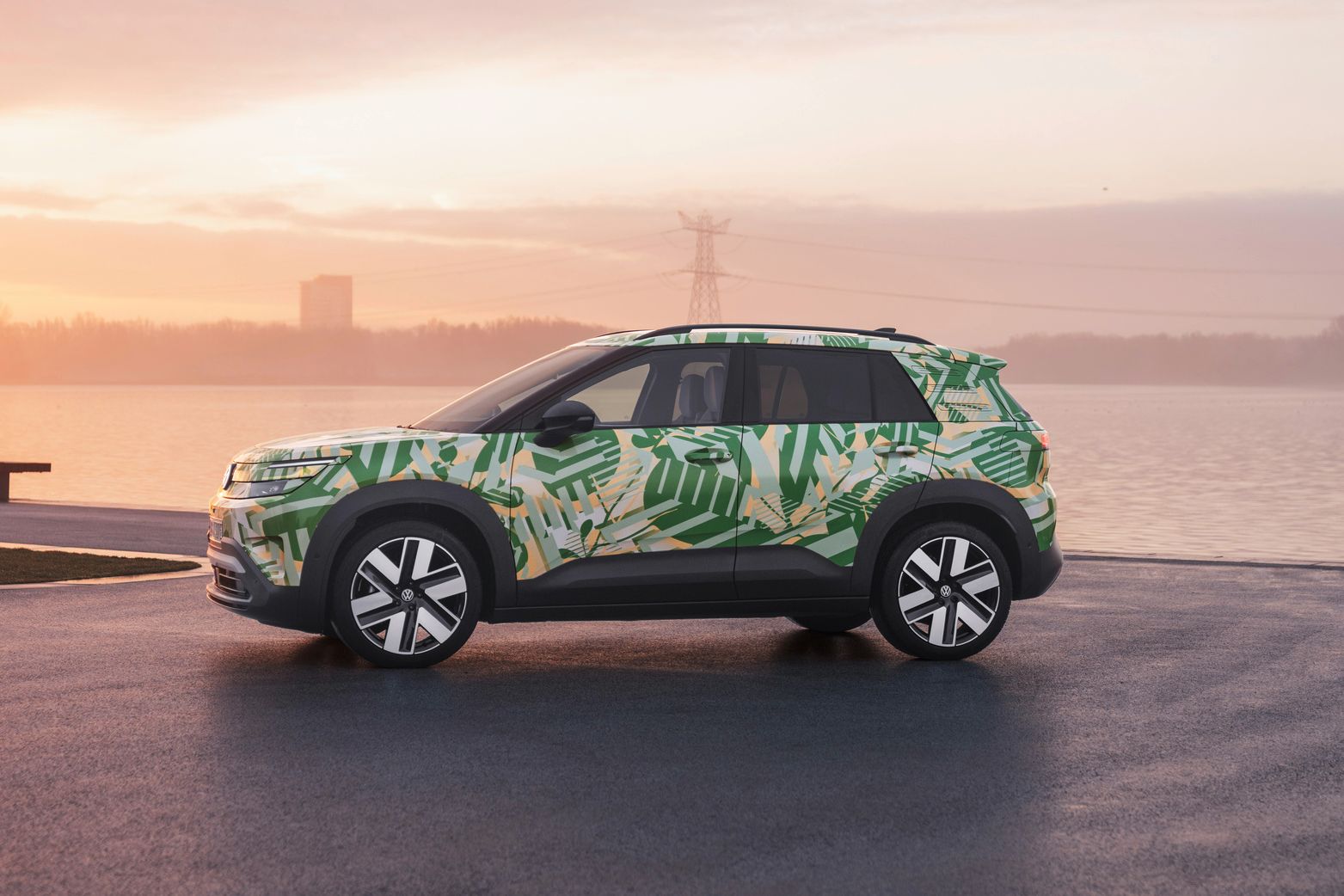 Ein SUV mit einer grünen und weißen Tarnlackierung steht auf einem asphaltierten Platz vor einem Gewässer bei Sonnenuntergang. Im Hintergrund sind Strommasten und Gebäude zu sehen. Der Himmel ist leicht bewölkt und in warmen Farben gehalten.