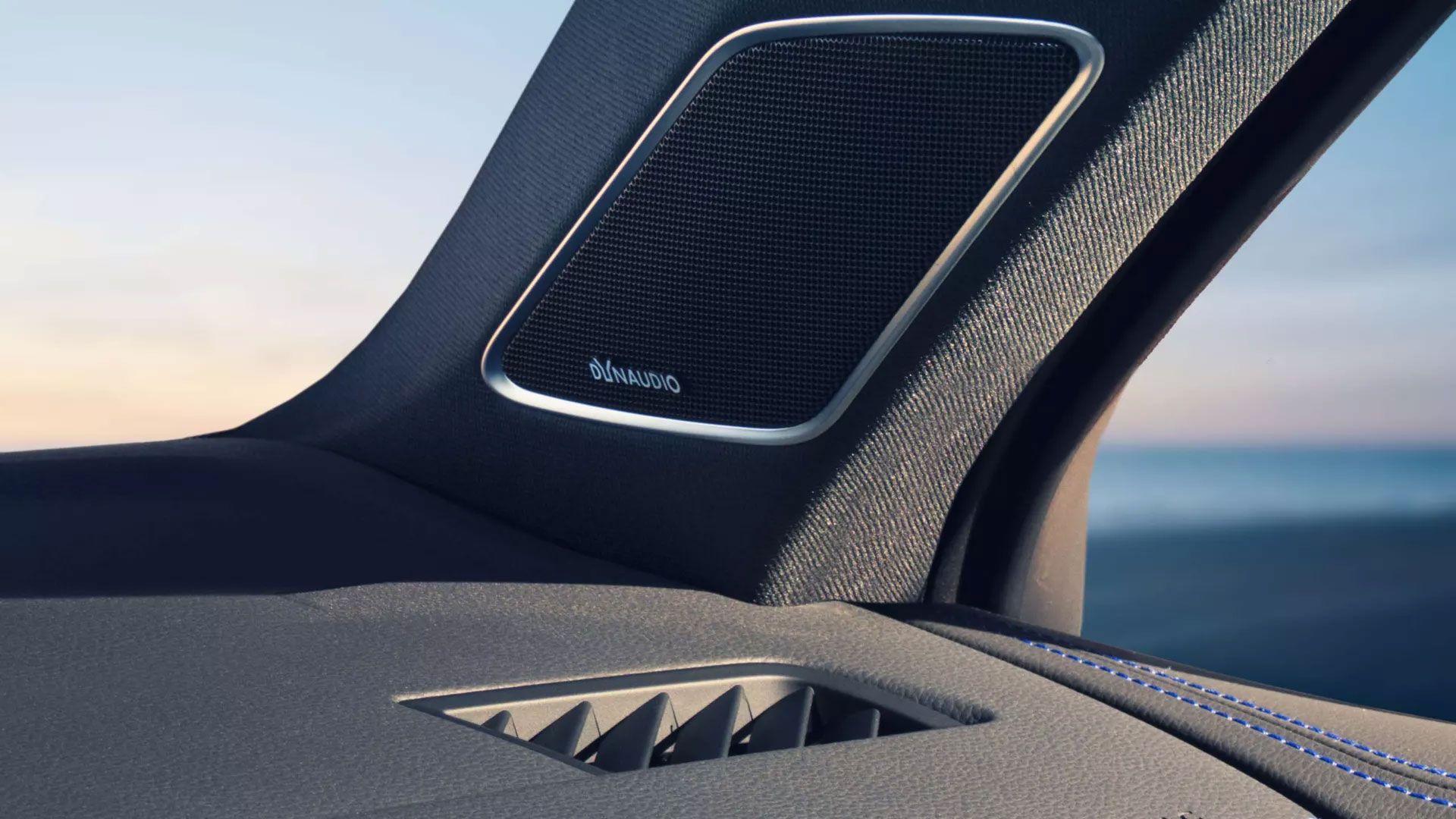 Nahaufnahme eines Lautsprechersystems im Innenraum eines  Touareg R . Der Lautsprecher zeigt den Schriftzug "DynAudio" und befindet sich auf einem stilvollen Textilpanel. Im Hintergrund ist durch das Fenster ein verschwommener Blick aufs Meer zu erkennen.