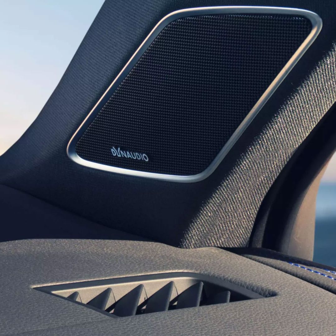 Nahaufnahme eines Lautsprechersystems im Innenraum eines  Touareg R . Der Lautsprecher zeigt den Schriftzug "DynAudio" und befindet sich auf einem stilvollen Textilpanel. Im Hintergrund ist durch das Fenster ein verschwommener Blick aufs Meer zu erkennen.