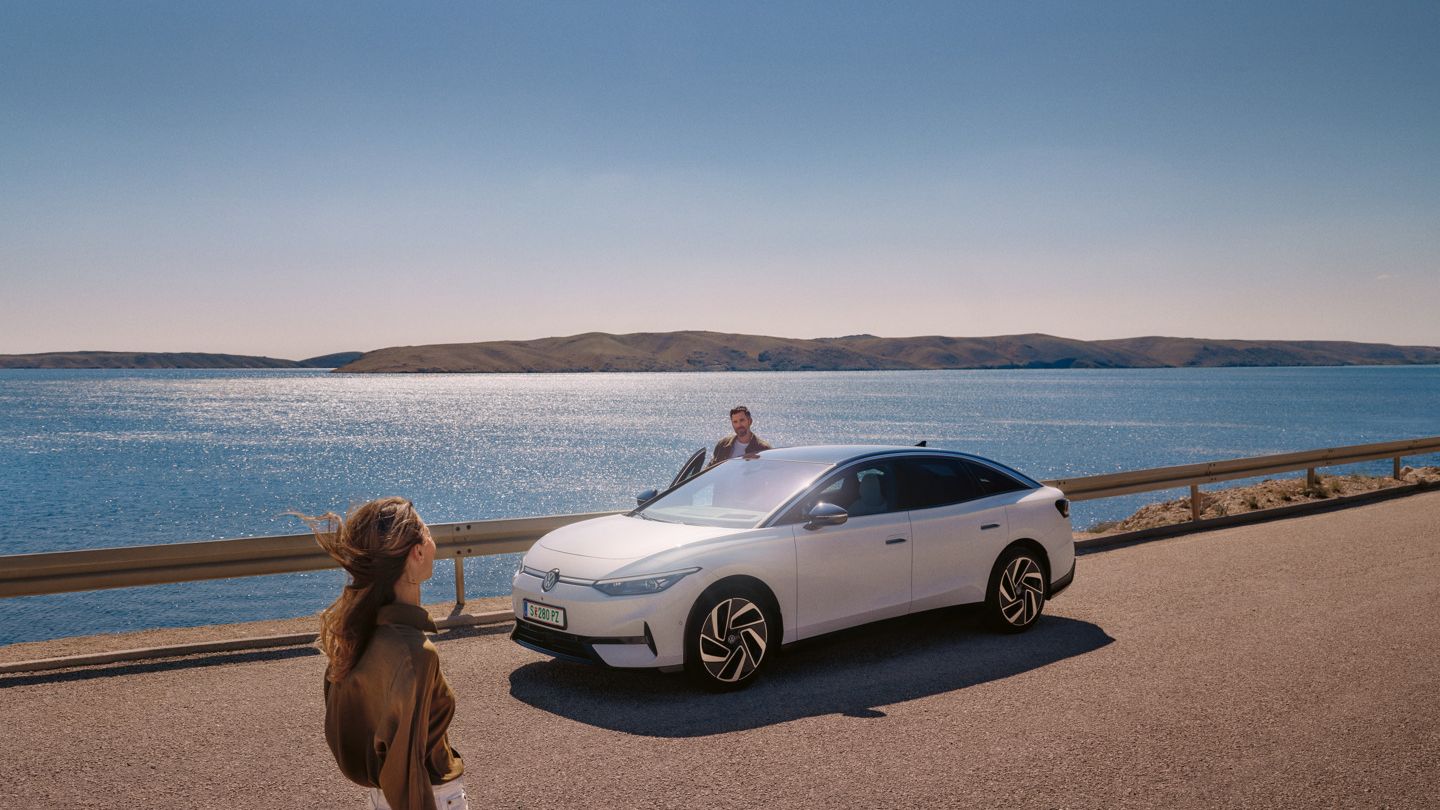 Eine Frau geht auf einen weißen VW ID.7 zu, der am Meer parkt. Auf der Beifahrerseite steigt ein Mann aus.