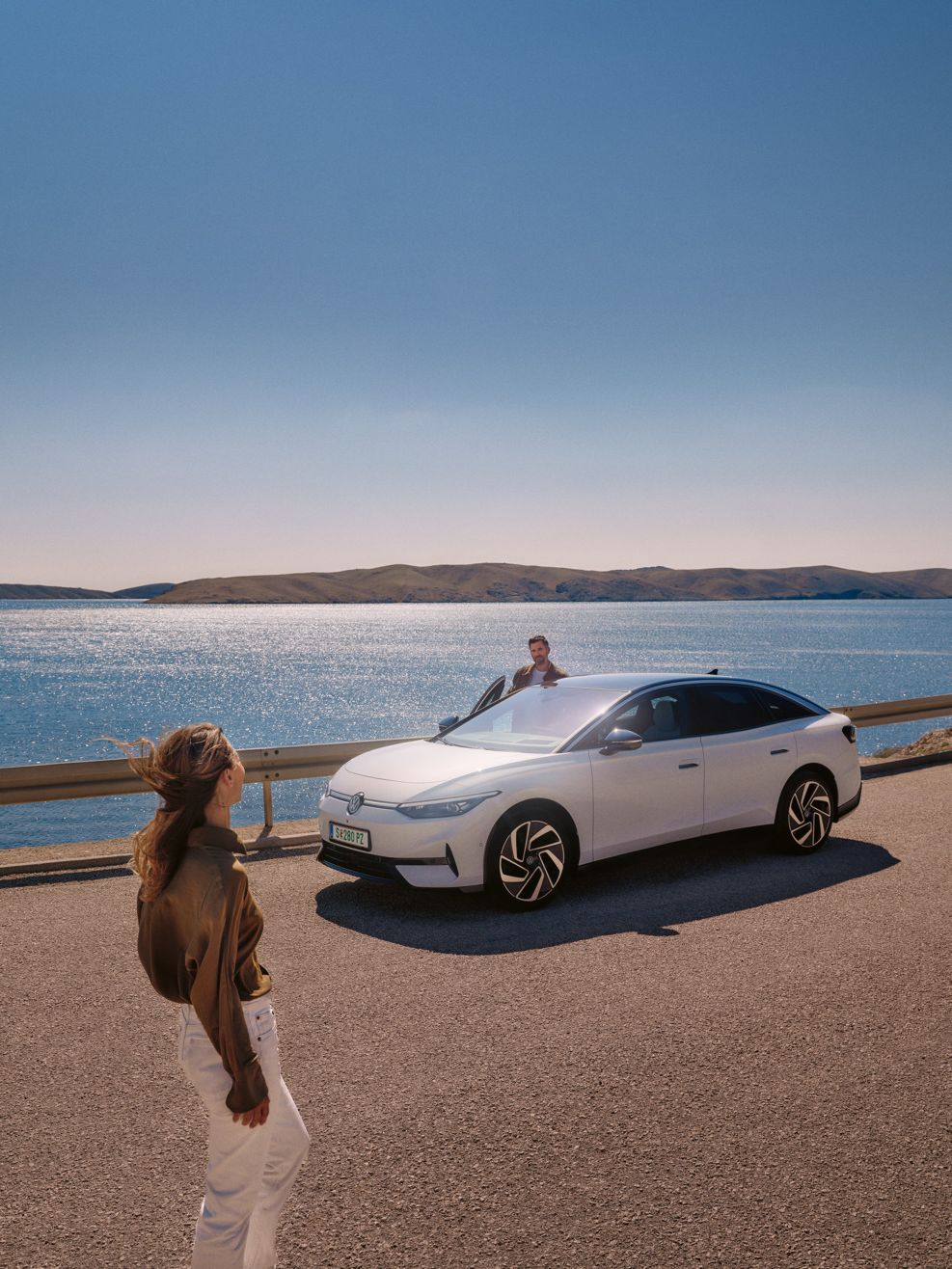 Eine Frau geht auf einen weißen VW ID.7 zu, der am Meer parkt. Auf der Beifahrerseite steigt ein Mann aus.