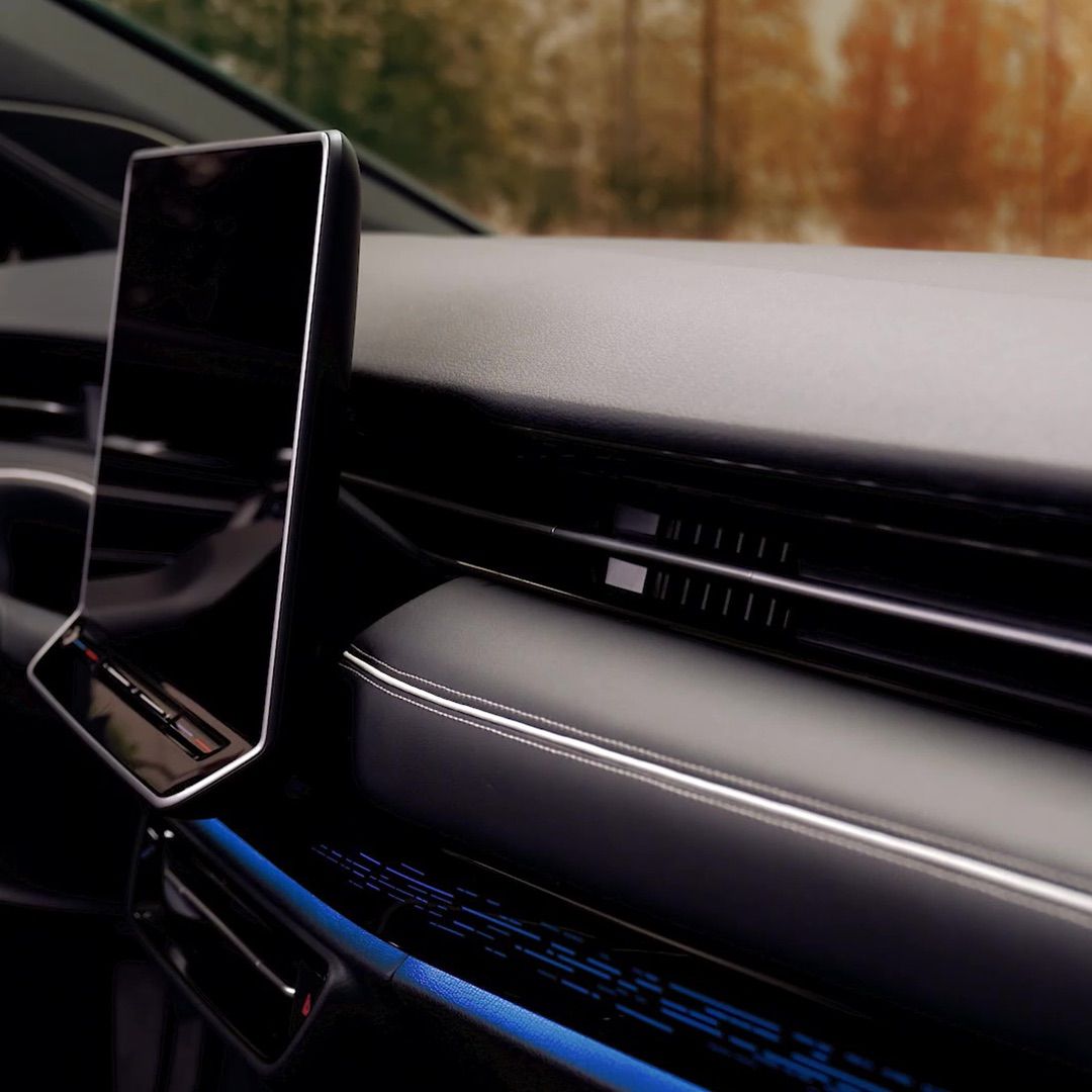 Nahaufnahme des Armaturenbretts eines ID.7 Tourer mit einem großen rechteckigen Display. Die Innenausstattung ist elegant und minimalistisch gestaltet. Im Hintergrund sind unscharf herbstliche Bäume zu sehen.
