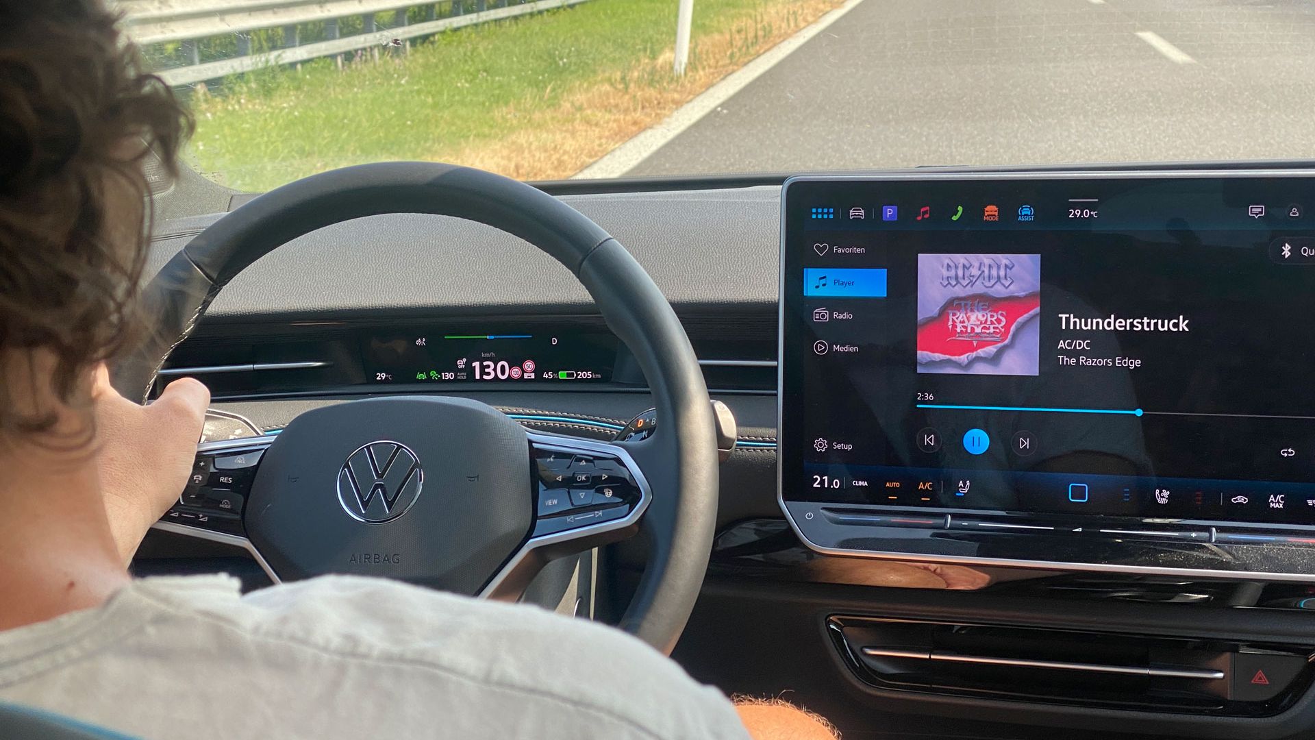 Eine Person fährt ein ID.7 und hält das Steuer mit beiden Händen. Auf dem Bildschirm des Autos wird das Lied "Thunderstruck" von AC/DC angezeigt. Die Straße vor dem Auto ist leer und das Wetter scheint klar zu sein.