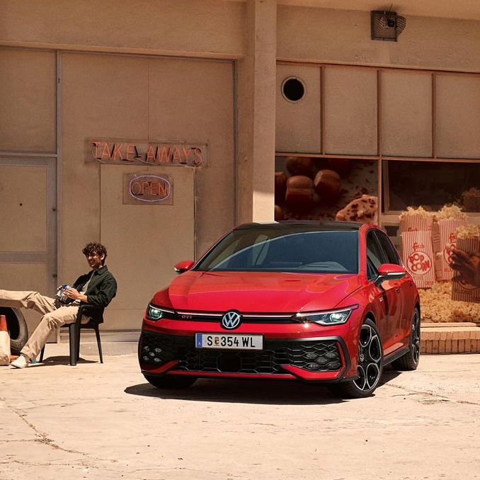 Zwei junge Männer sitzen vor einem Geschäft neben einem roten VW Golf GTI.