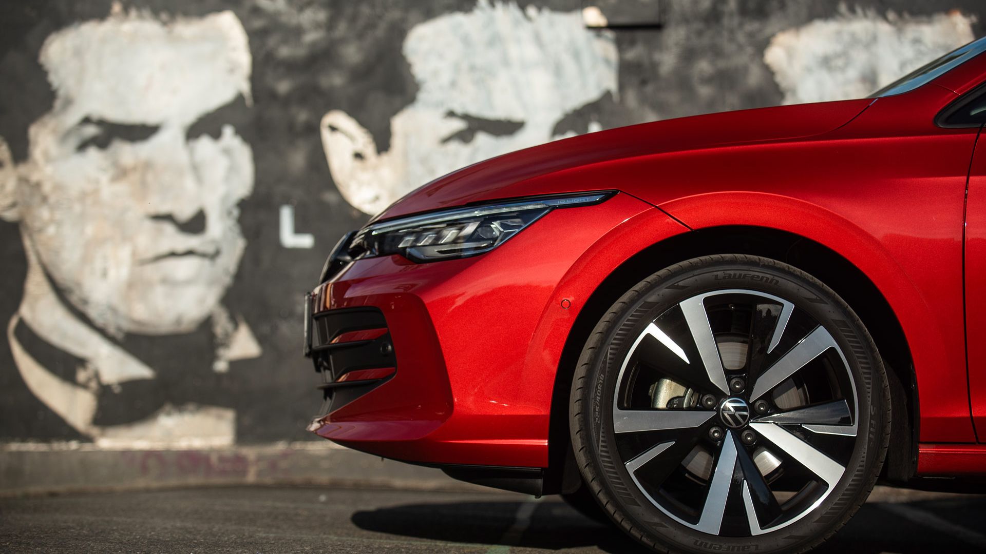 Seitenansicht der Front eines roten Autos, im Fokus der rechte Vorderreifen mit sportlicher Felgenoptik. Im Hintergrund ist eine dunkle, leicht verwitterte Wand mit übermalten Graffitis zu sehen. Links im Bild ist ein rechteckiger, unscharf abgedeckter Bereich.