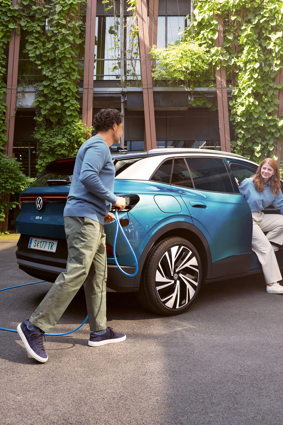 Eine Frau steigt aus einem blauem ID.4 aus, während ein Mann das POWER 2 Go Kabel anschließt. Im Hintergrund sind grüne Pflanzen und moderne Gebäude zu sehen.