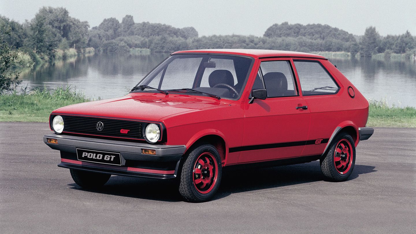 Ein roter Volkswagen Polo GT steht auf einem Asphalt vor einer natürlichen Landschaft mit See und Bäumen im Hintergrund. Das Auto verfügt über schwarzen Akzente und rote Felgen. Es ist ein sportliches Modell mit zwei Türen.