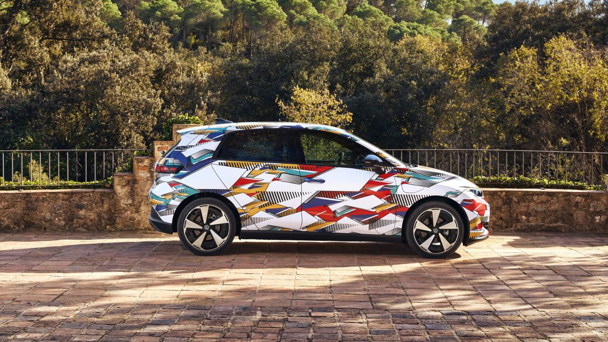 Ein ID. Polo mit buntem, geometrisch gemustertem Aufdruck steht auf einem gepflasterten Platz. Hinter dem Auto erstreckt sich eine bewaldete, hügelige Landschaft. Die Sonne scheint, und das Wetter ist klar.
