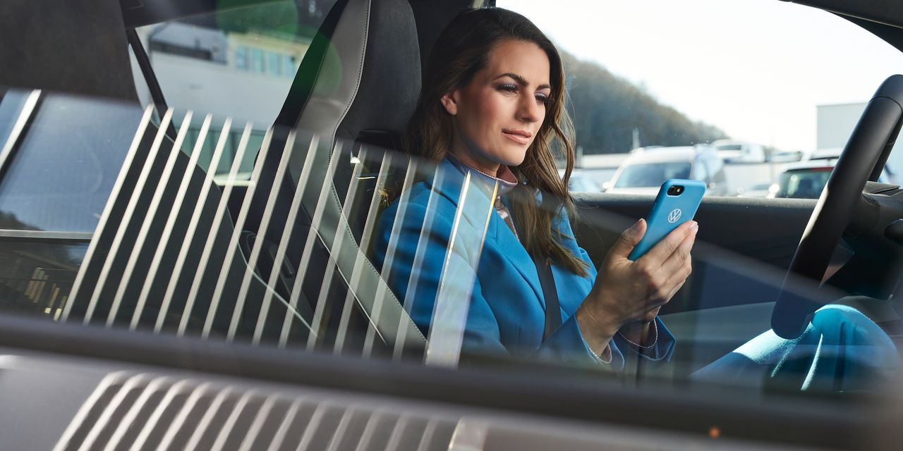 Eine in blau gekleidete Frau sitzt in einem geparkten Volkswagen mit einem Smartphone in ihrer Hand.