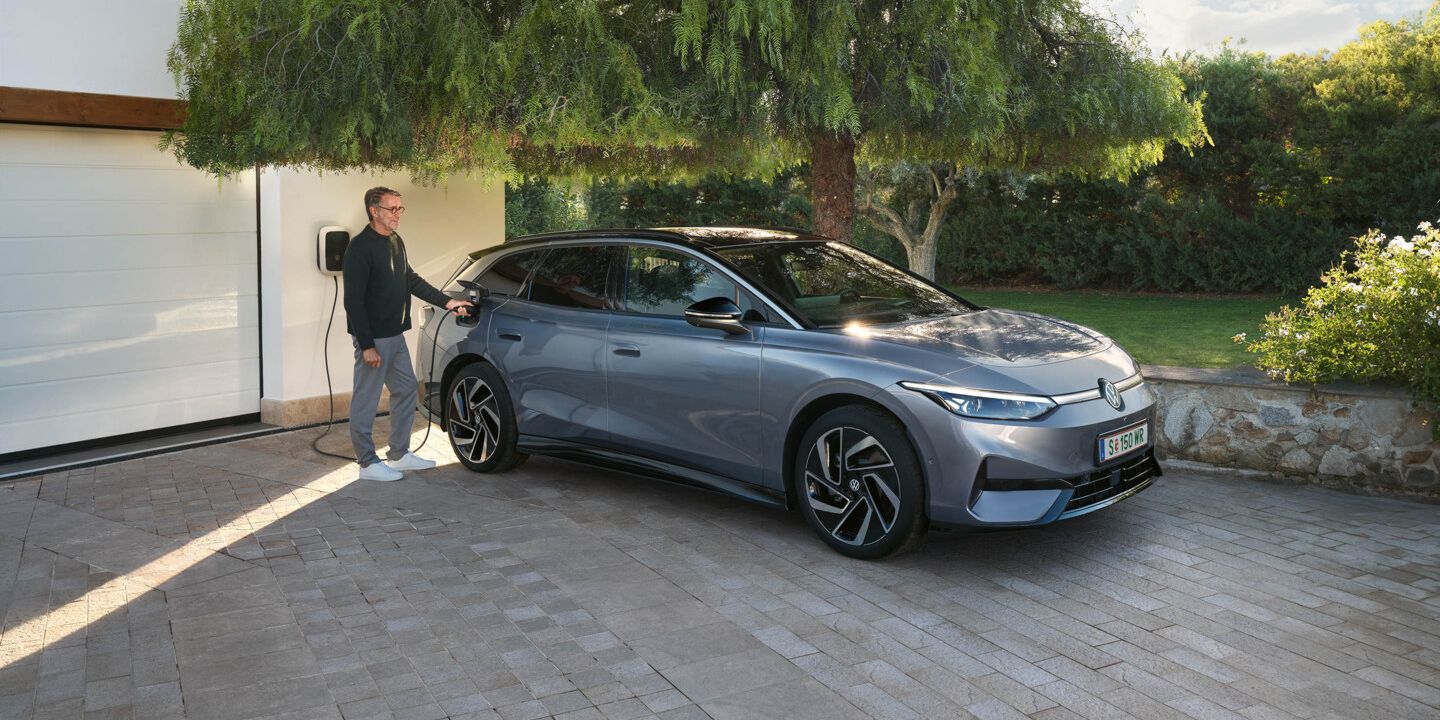 Ein ID.7 Tourer steht in einer Einfahrt vor einem Haus, ein Mann steckt den Ladestecker in die Ladebuchse. 