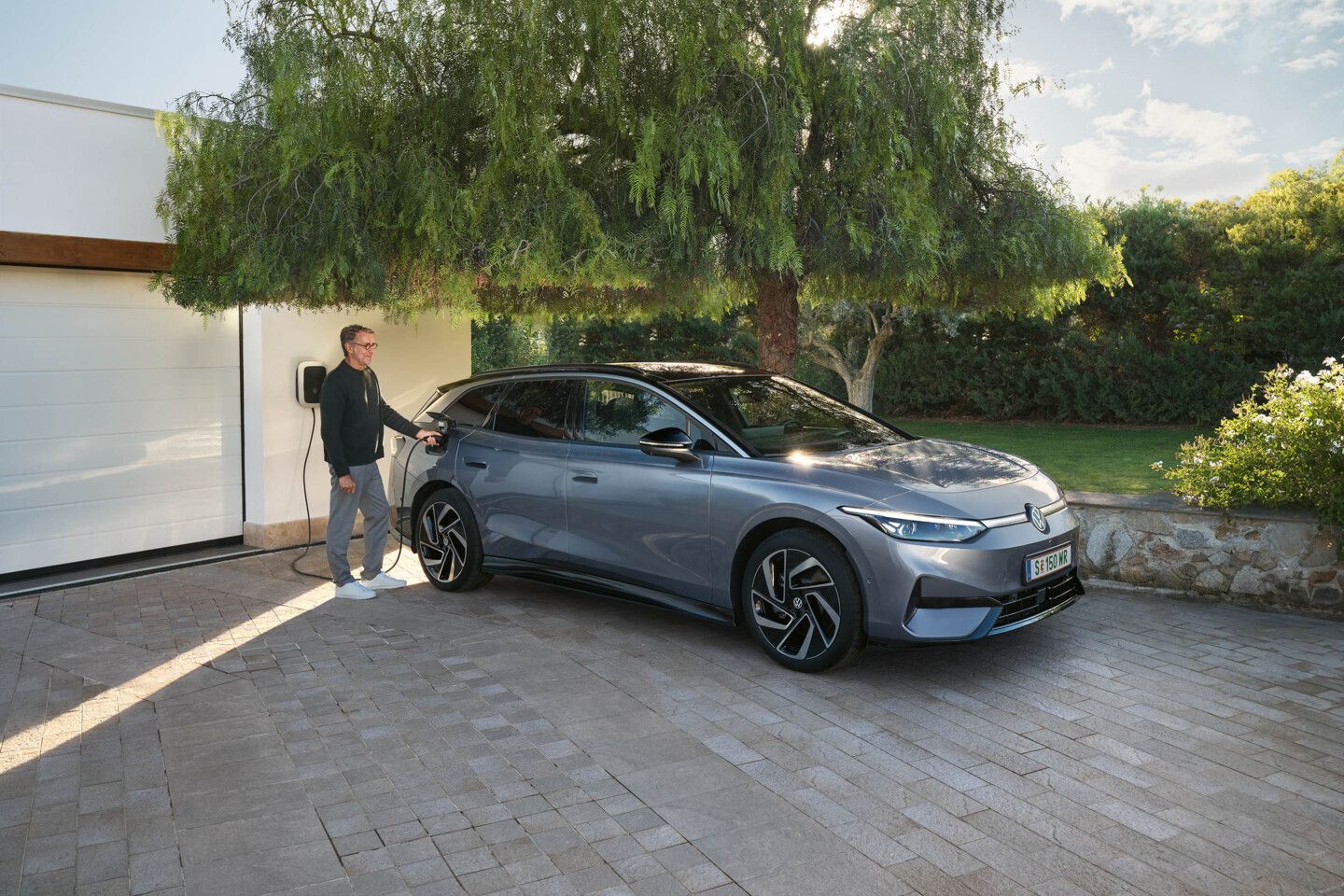 Ein ID.7 Tourer steht in einer Einfahrt vor einem Haus, ein Mann steckt den Ladestecker in die Ladebuchse. 