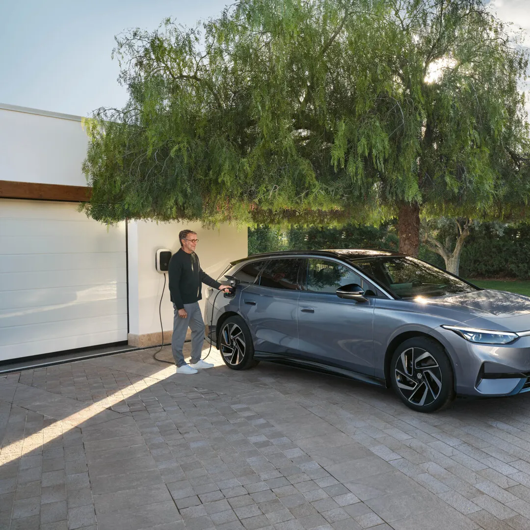 Ein ID.7 Tourer steht in einer Einfahrt vor einem Haus, ein Mann steckt den Ladestecker in die Ladebuchse. 