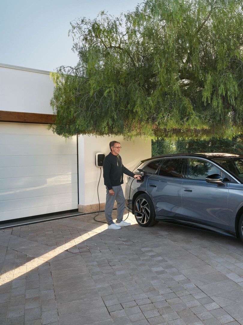 Ein ID.7 Tourer steht in einer Einfahrt vor einem Haus, ein Mann steckt den Ladestecker in die Ladebuchse. 