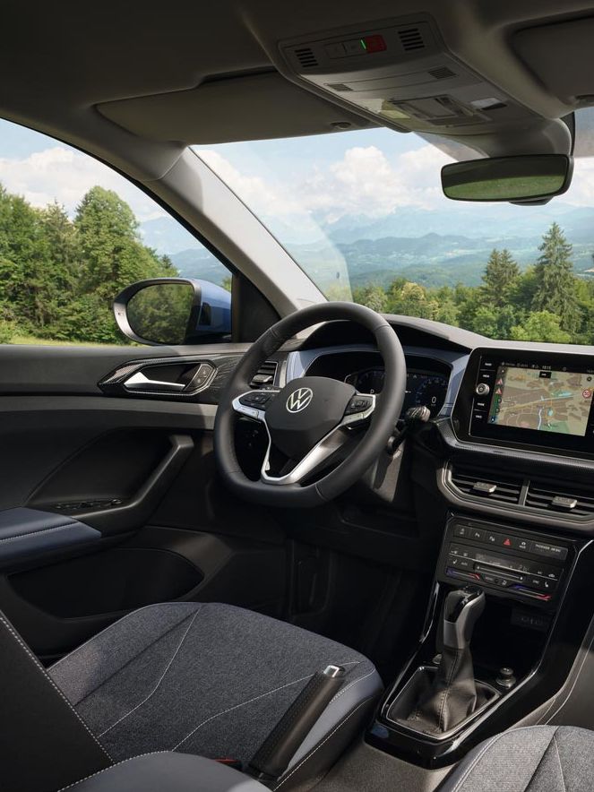 Blick vom Beifahrersitz eines VW T-Cross durch die Frontscheibe auf ein lachendes Pärchen vor Wald und Bergen.