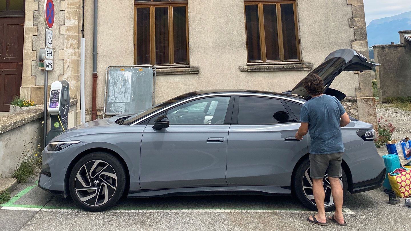 Ein Mann steht neben einem ID.7, das an einer Ladestation angeschlossen ist. Der Kofferraum des Autos ist geöffnet. Das Gebäude im Hintergrund hat große Fenster.