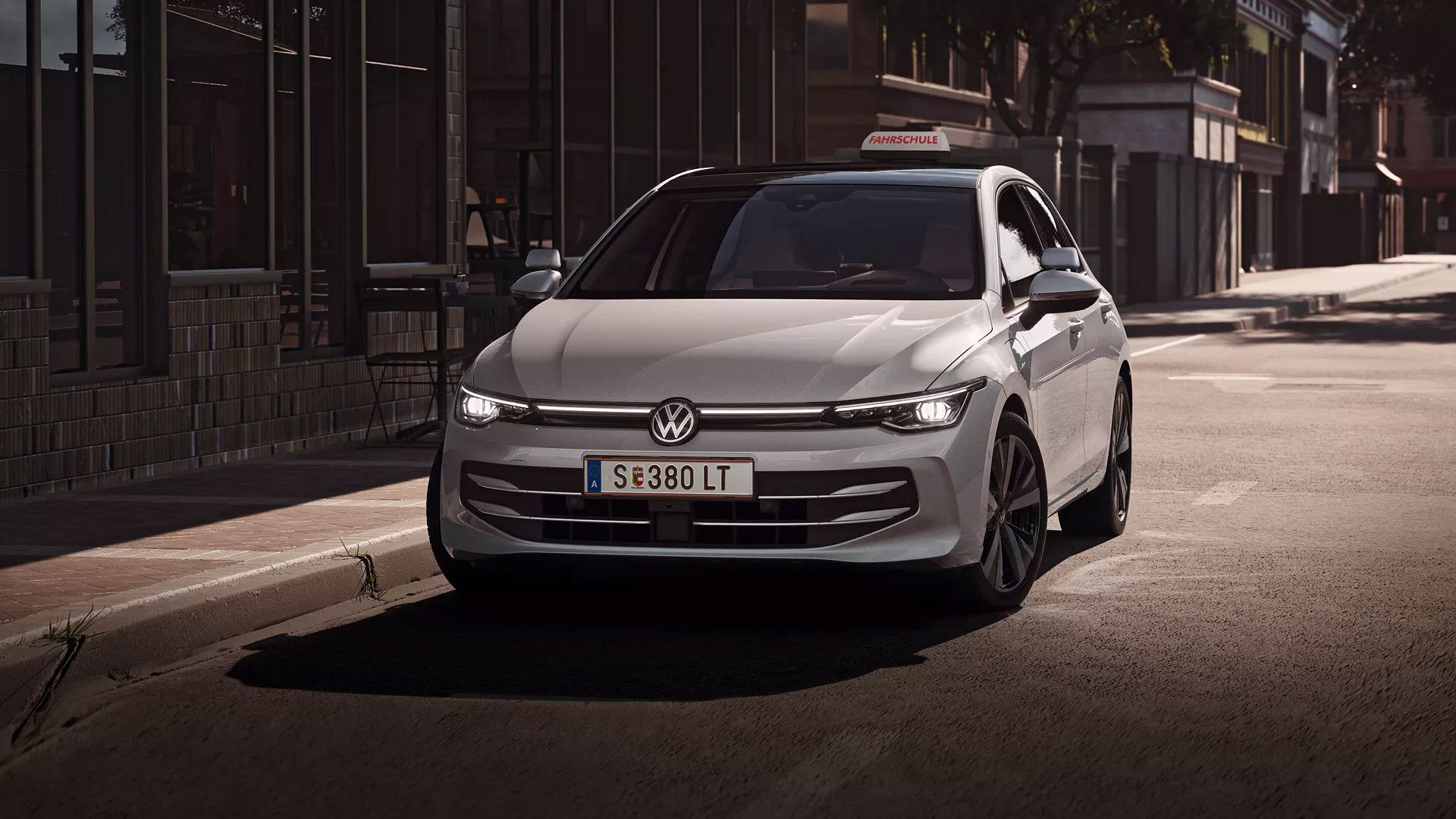 Ein weißes Volkswagen-Fahrzeug parkt auf einer städtischen Straße. Auf dem Fahrzeugdach ist ein Fahrer-Trainingsschild angebracht. Im Hintergrund sind Gebäude und Bäume zu sehen.