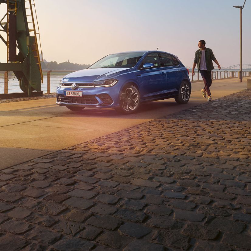  Ein blauer VW Polo R-Line steht an der Promenade neben einem Fluss, ein Mann läuft um das Auto herum.