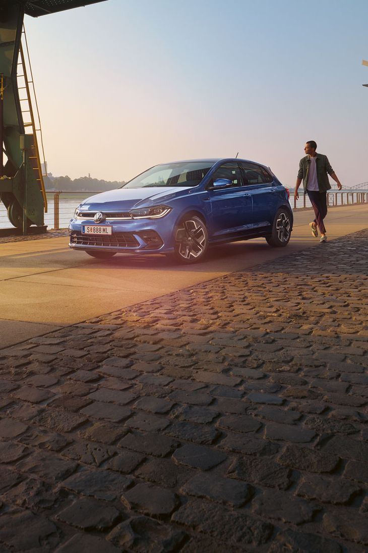  Ein blauer VW Polo R-Line steht an der Promenade neben einem Fluss, ein Mann läuft um das Auto herum.