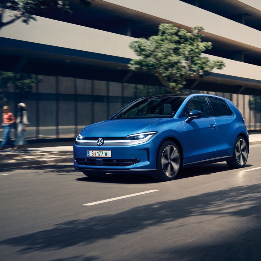 Ein blauer VW ID. Polo Style fährt durch eine Stadt mit grünen Bäumen am Straßenrand.