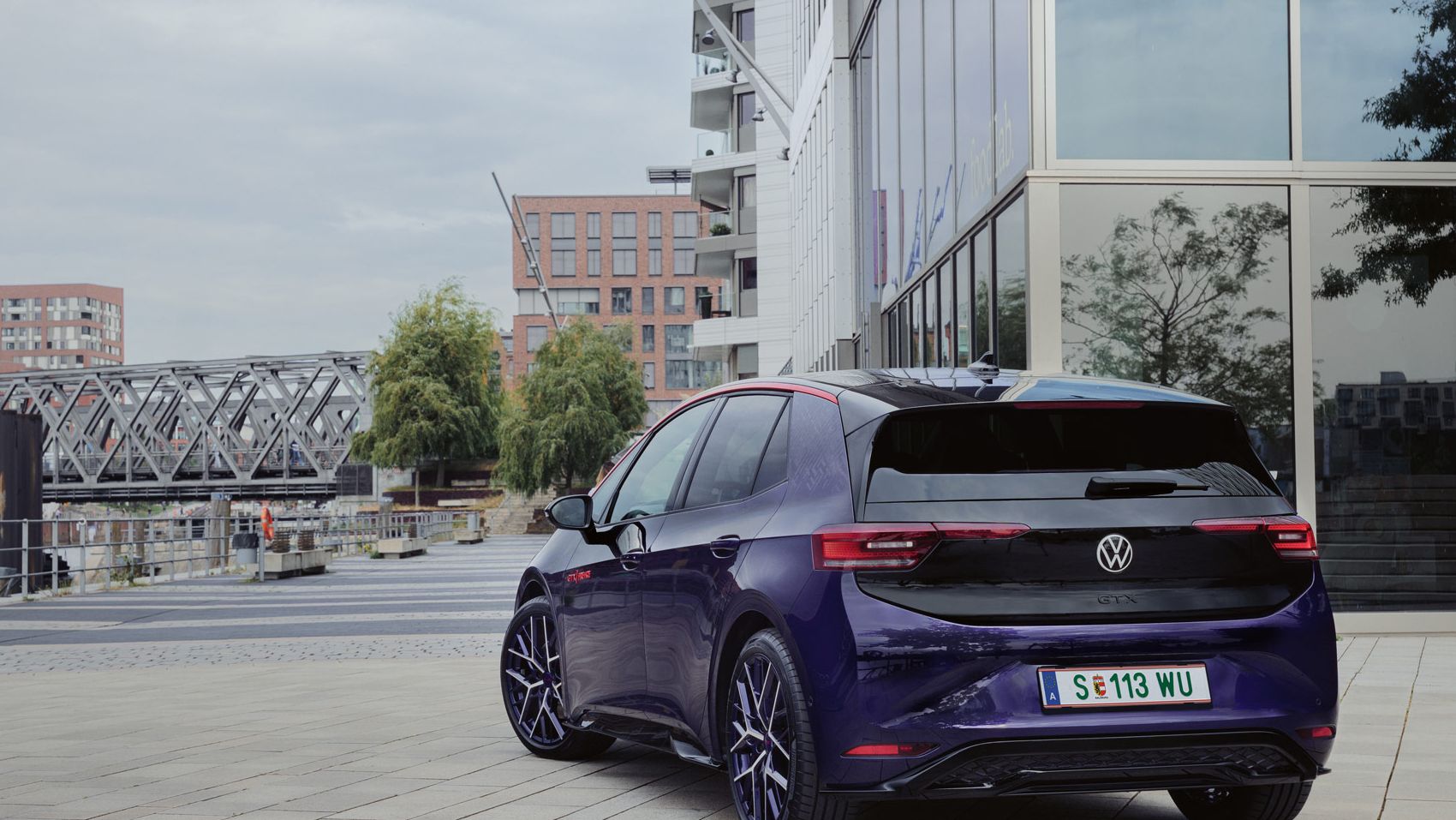 Elektrisches Volkswagen Auto geparkt auf einem modernen Stadtplatz. Im Hintergrund sind Gebäude und eine Brücke zu sehen. Das Auto hat ein Kennzeichen aus Wolfsburg.
