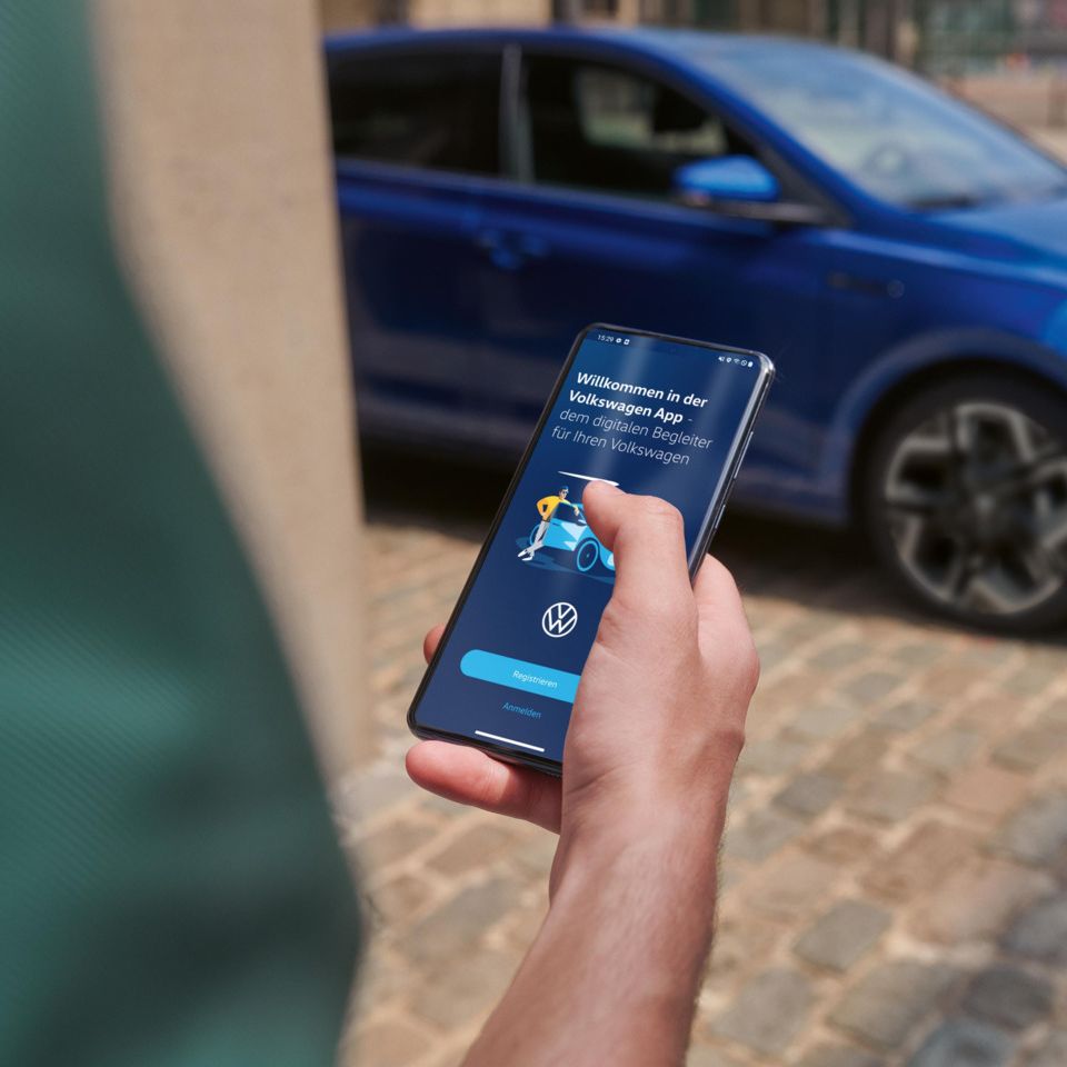 Eine Person hält ein Smartphone in der Hand mit der Volkswagen App, im Hintergrund steht ein blauer VW Polo R-Line.