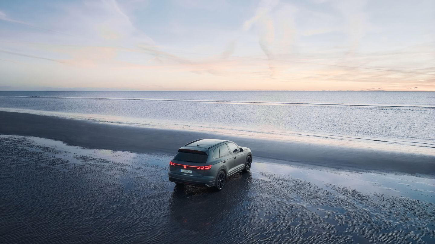 Ein Taoureg R steht auf nassem Sandstrand bei Sonnenuntergang. Das Meer ist im Hintergrund zu sehen, der Himmel ist leicht bewölkt. Die Rücklichter des Autos sind hell erleuchtet.