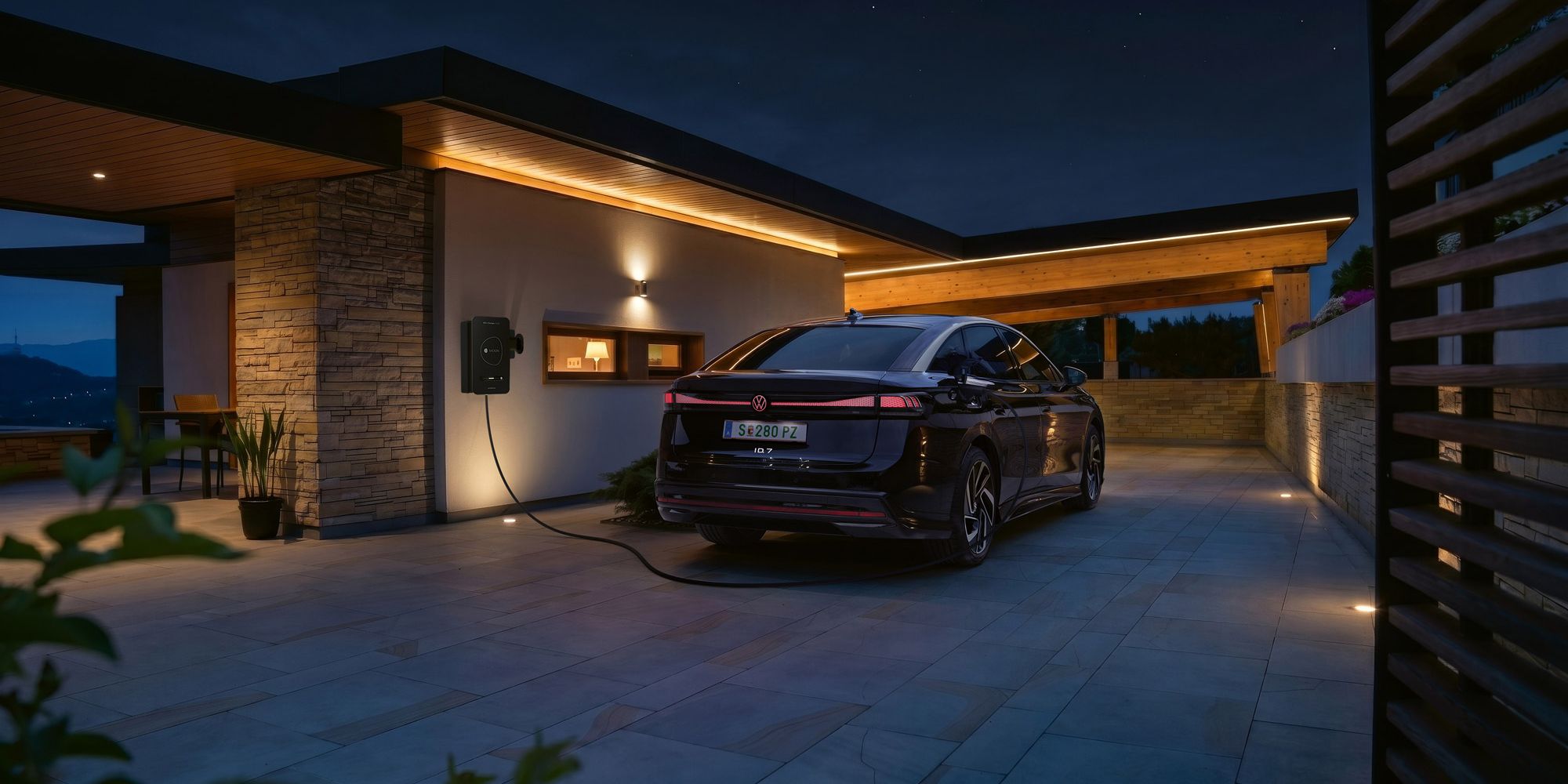 Ein schwarzes Elektroauto steht nachts in der Einfahrt eines modernen Hauses und ist an einer Wallbox zum Laden angeschlossen. Warmes, indirektes Licht an Fassade und Carport beleuchtet die Szene stimmungsvoll. Im Hintergrund sind Berge oder Hügel im Dämmerlicht zu erkennen.