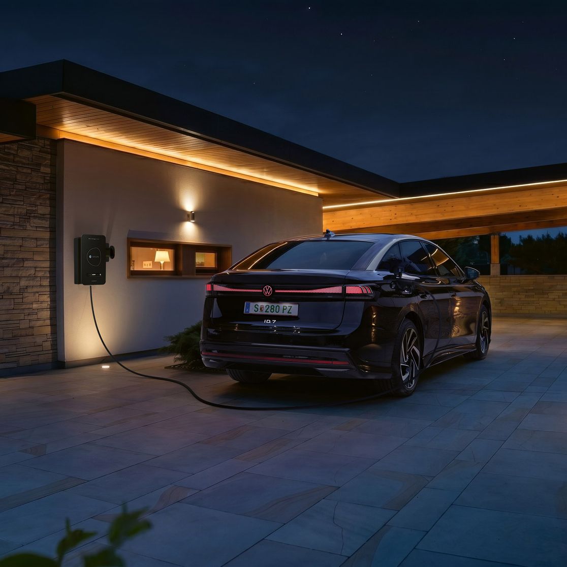 Ein schwarzes Elektroauto steht nachts in der Einfahrt eines modernen Hauses und ist an einer Wallbox zum Laden angeschlossen. Warmes, indirektes Licht an Fassade und Carport beleuchtet die Szene stimmungsvoll. Im Hintergrund sind Berge oder Hügel im Dämmerlicht zu erkennen.