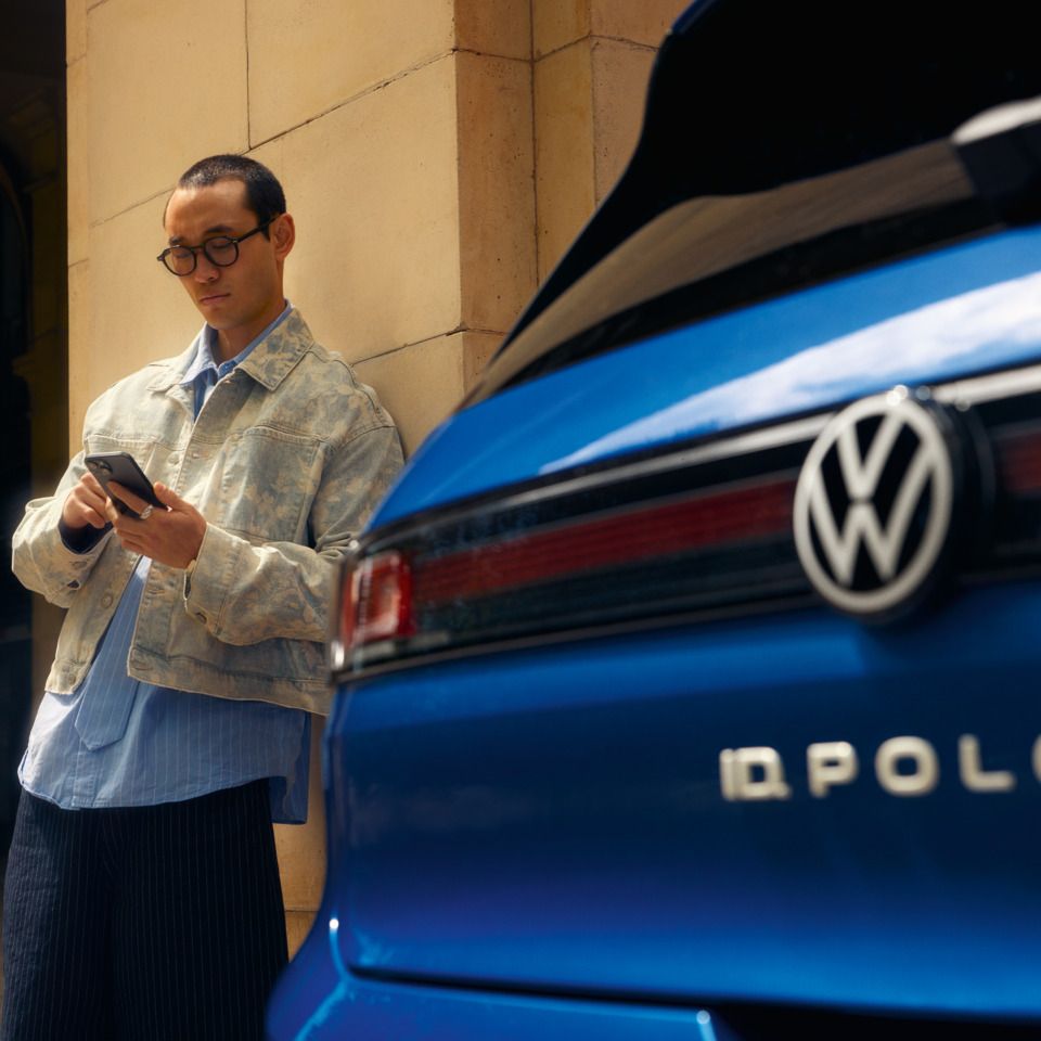 Heckansicht eines blauen VW ID. Polo, daneben bedient ein Mann sein Handy.