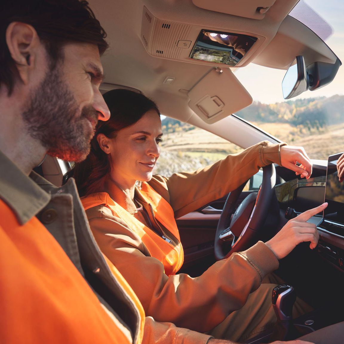 Zwei Personen in orangefarbenen Warnwesten sitzen in einem Auto und betrachten den Bildschirm des Infotainmentsystems. 