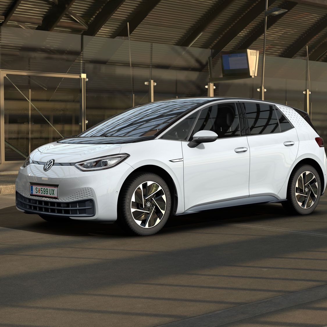 Ein weißer Volkswagen ID.3 steht in einer modernen, teilweise verglasten Tiefgarage. Das Auto ist von der Seite und leicht von vorne zu sehen. Im Hintergrund sind Maschendrahtzäune und Betonwände zu erkennen.