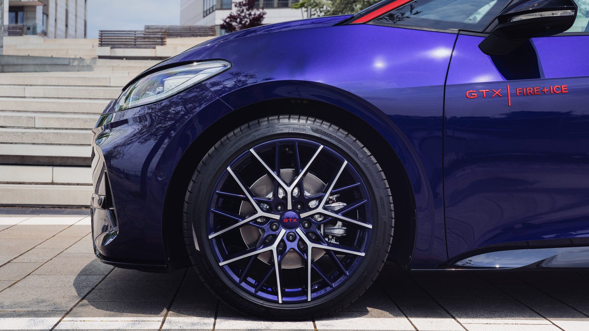 Nahaufnahme eines Autoreifens mit auffälligen blau-lila Felgen. Das Auto hat eine lilafarbene Karosserie und die Aufschrift "GTX FIRE+ICE". Im Hintergrund sind moderne Gebäude und Treppen zu sehen.
