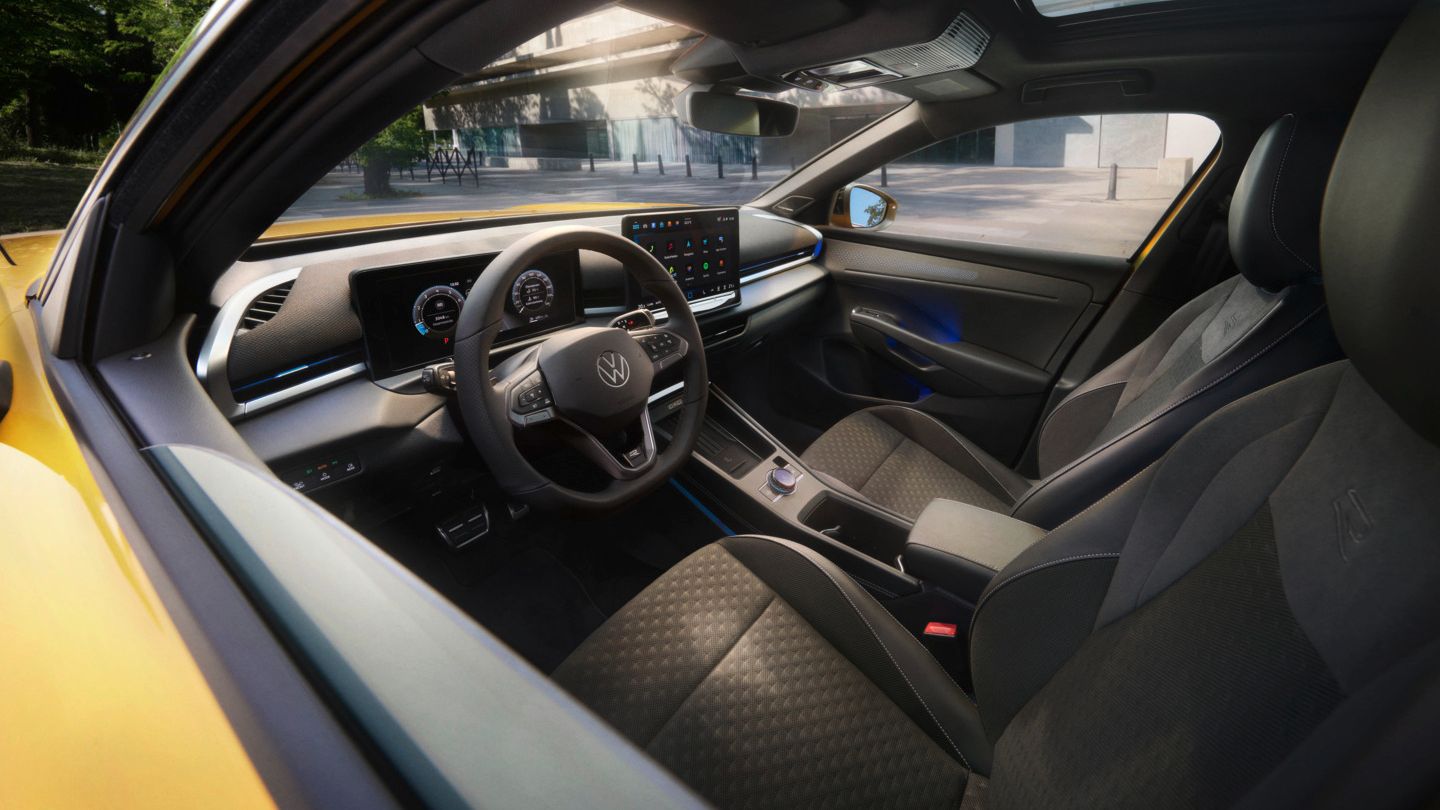 VW T-Roc Das bild zeigt den Blick in das Interieur eines gelben VW T-Rocs. Man sieht die beiden vorderen Sitze, ein Lenkrad, das Infotainmentsystem aber auch einen kleinen Teil des gelb lackierten Exterieurs. Im Hintergrund sieht man Ein städtisches Gebäude und daneben dichte Laubbäume.