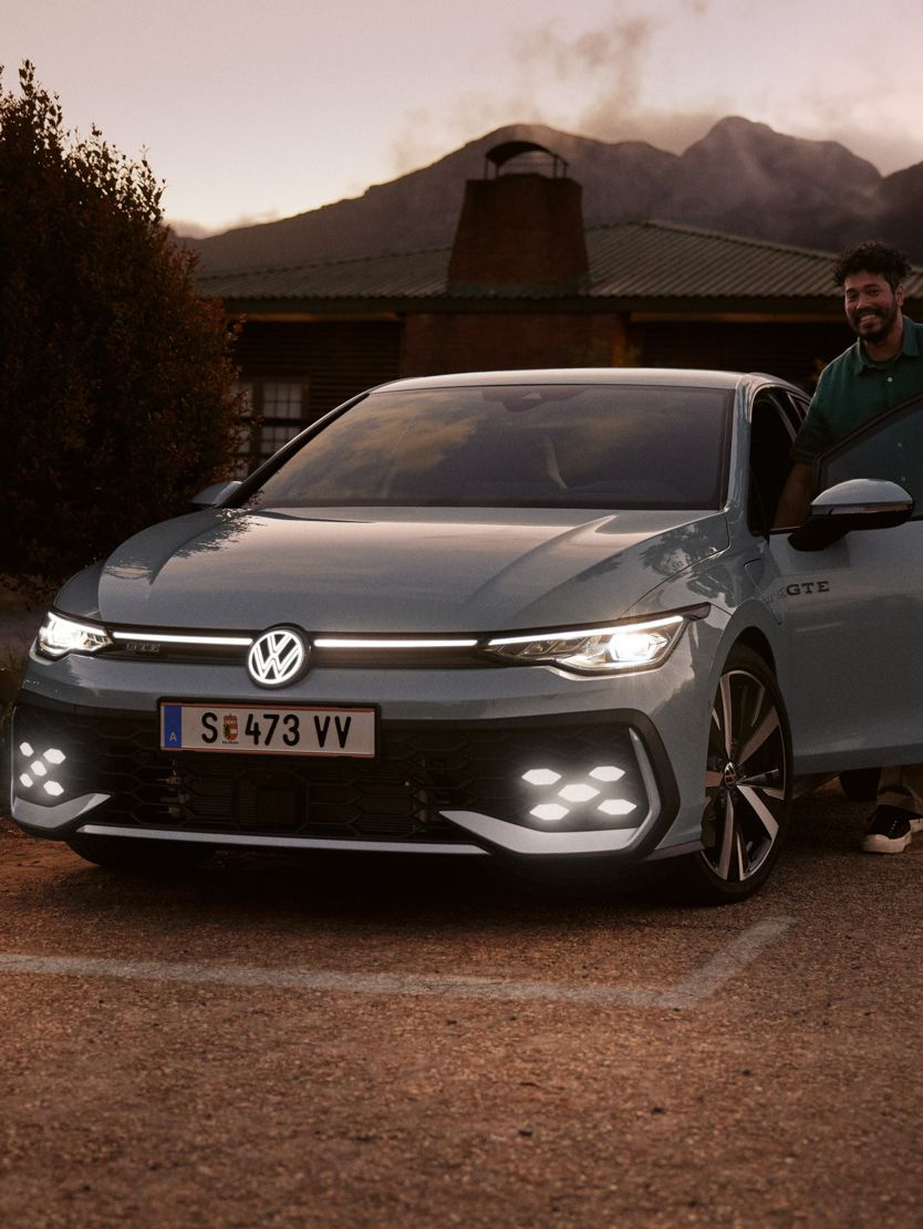 Frontansicht eines grauen VW Golf GTE mit IQ. Light LED Matrix Scheinwerfern in abendlicher Stimmung, Mann steigt in Golf GTE