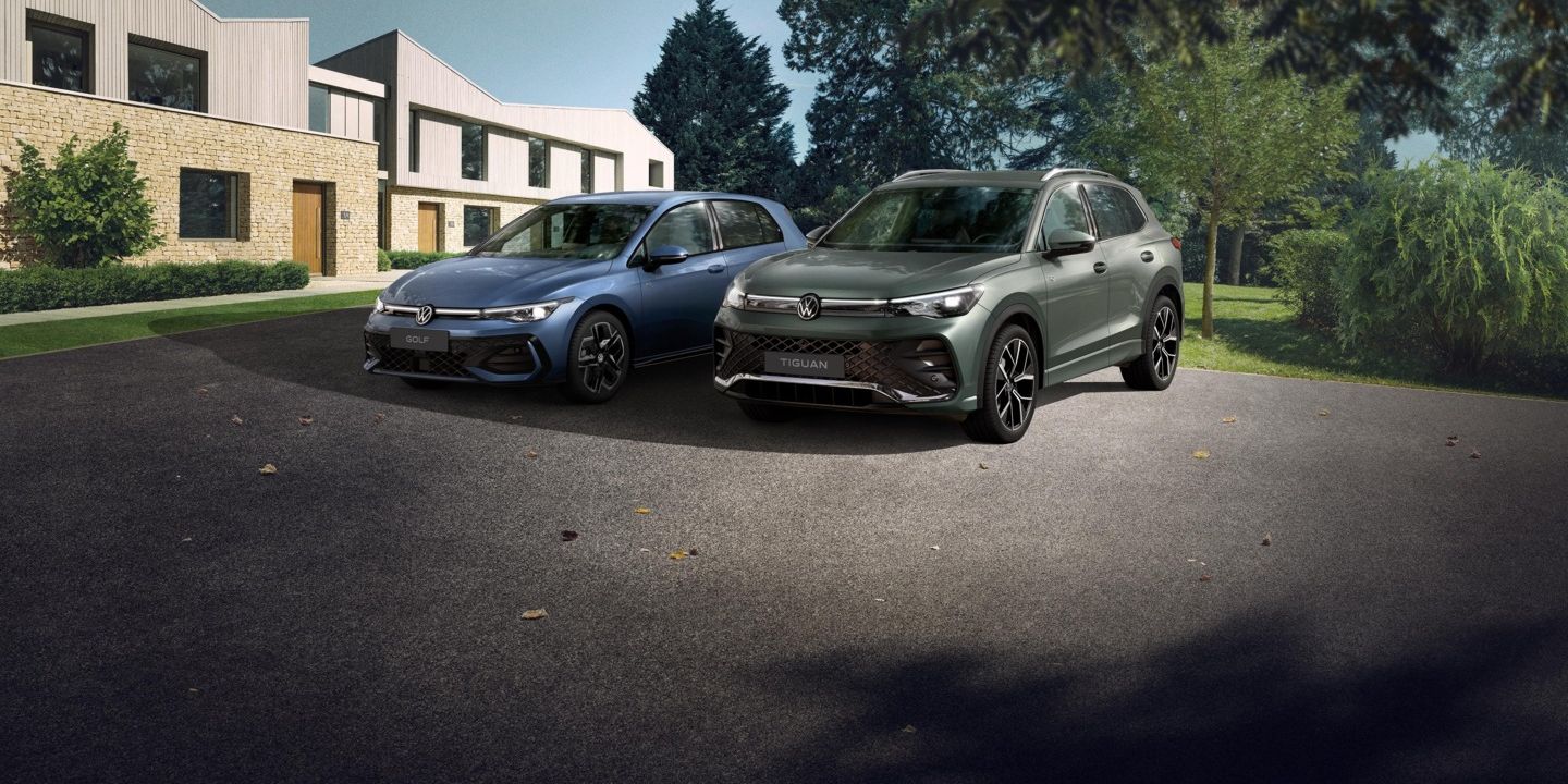 Zwei VW Modell, ein blauer Golf und ein grüner Tiguan, stehen nebeneinander auf einer Einfahrt vor einem Reihenhaus. Das Umfeld ist grün und bewaldet, das Wetter scheint klar zu sein.